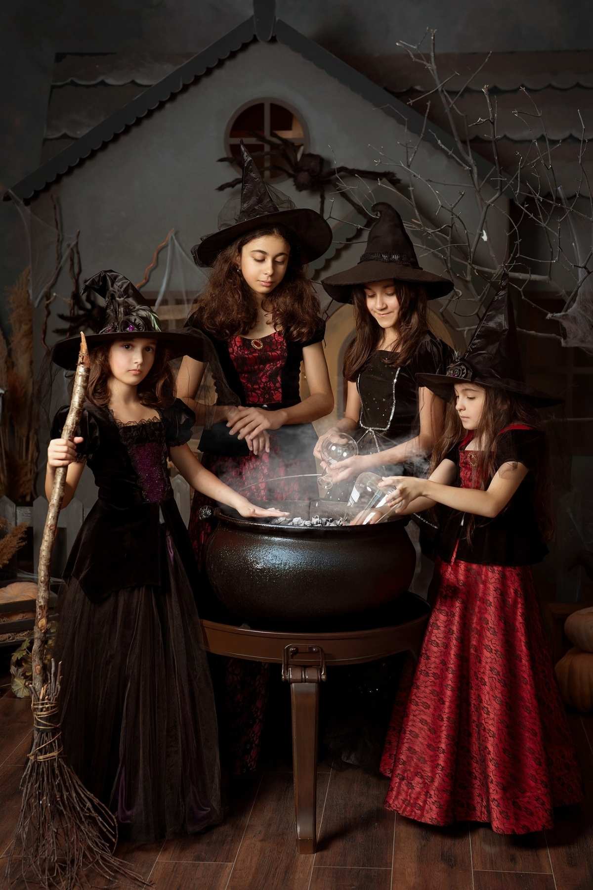 Four girls in witch costumes stand around a steaming cauldron, with a broom on the left and a spooky house backdrop behind them.