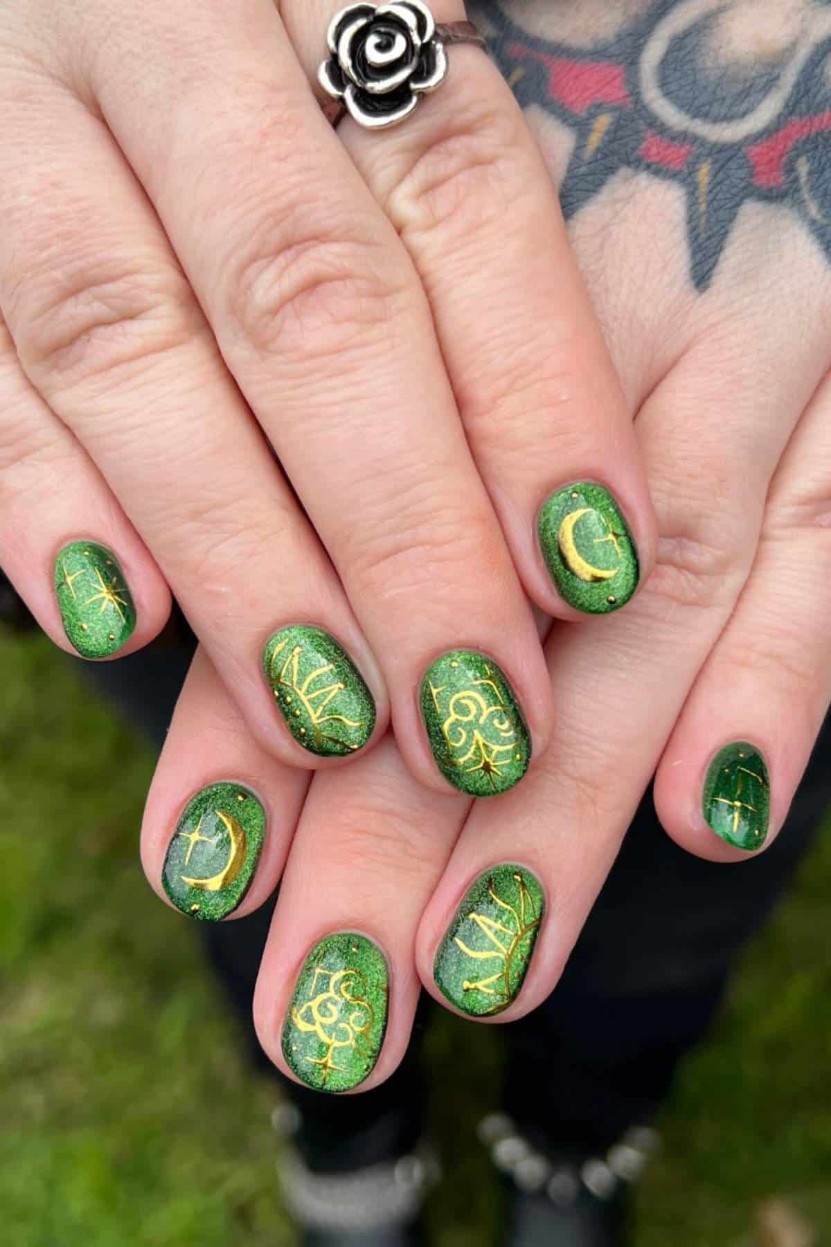 Hands with green nails featuring gold crescent moons, dots, and vine designs. Person wears a silver rose ring and has a colorful tattoo on their wrist. Grass is visible in the background.