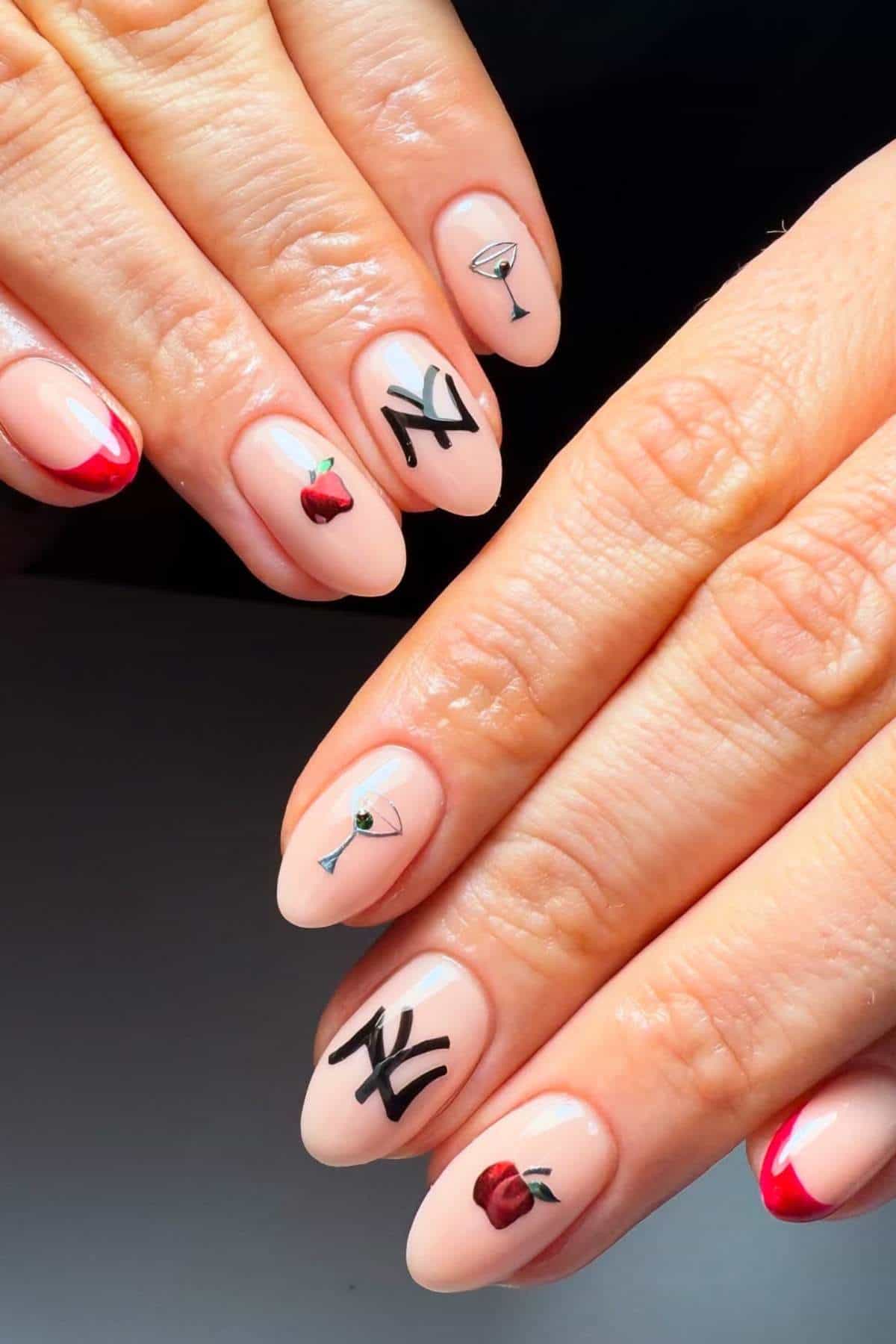 Hands with nude almond-shaped nails featuring nail art designs: red tips, black "NY" letters, a red apple, and a martini glass on different nails.