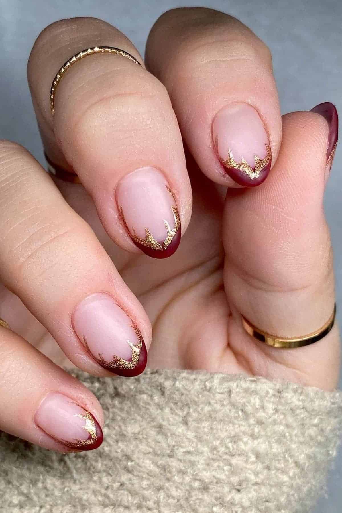 A hand with short, rounded nails featuring burgundy French tips and gold antler accents. The person wears gold rings and a beige knitted sleeve is visible.