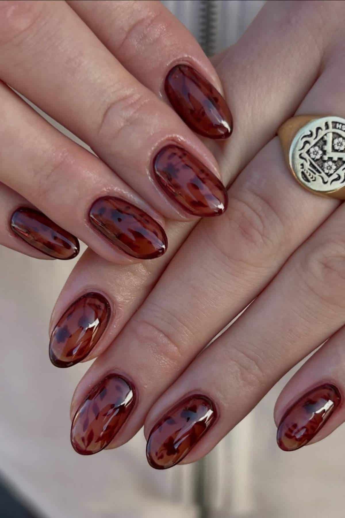 Close-up of hands with almond-shaped nails featuring a glossy, dark tortoiseshell pattern. A gold ring with an engraved design is worn on one finger.