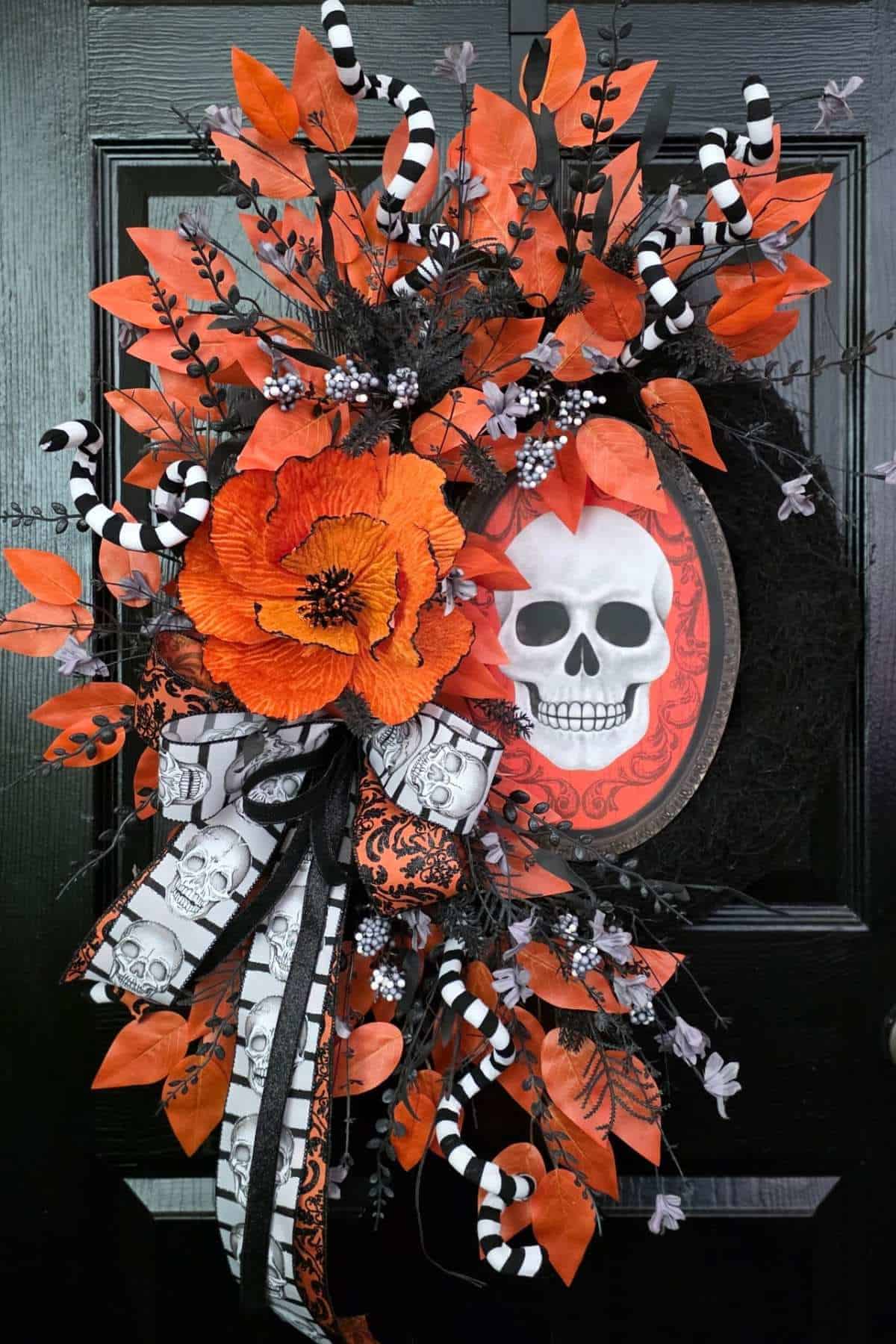 Wreath with orange leaves, black branches, striped accents, large orange flower, and framed skull art hangs on a black door.