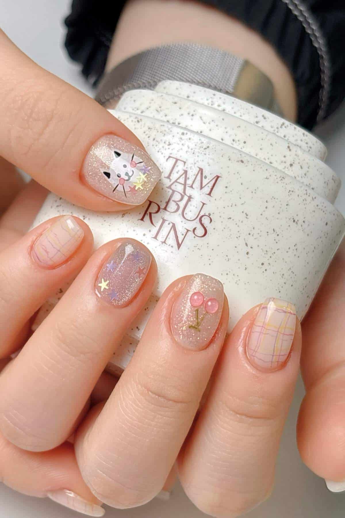 A hand with pastel nail art featuring a cat face, cherry design, stars, and plaid patterns, holding a white speckled container.