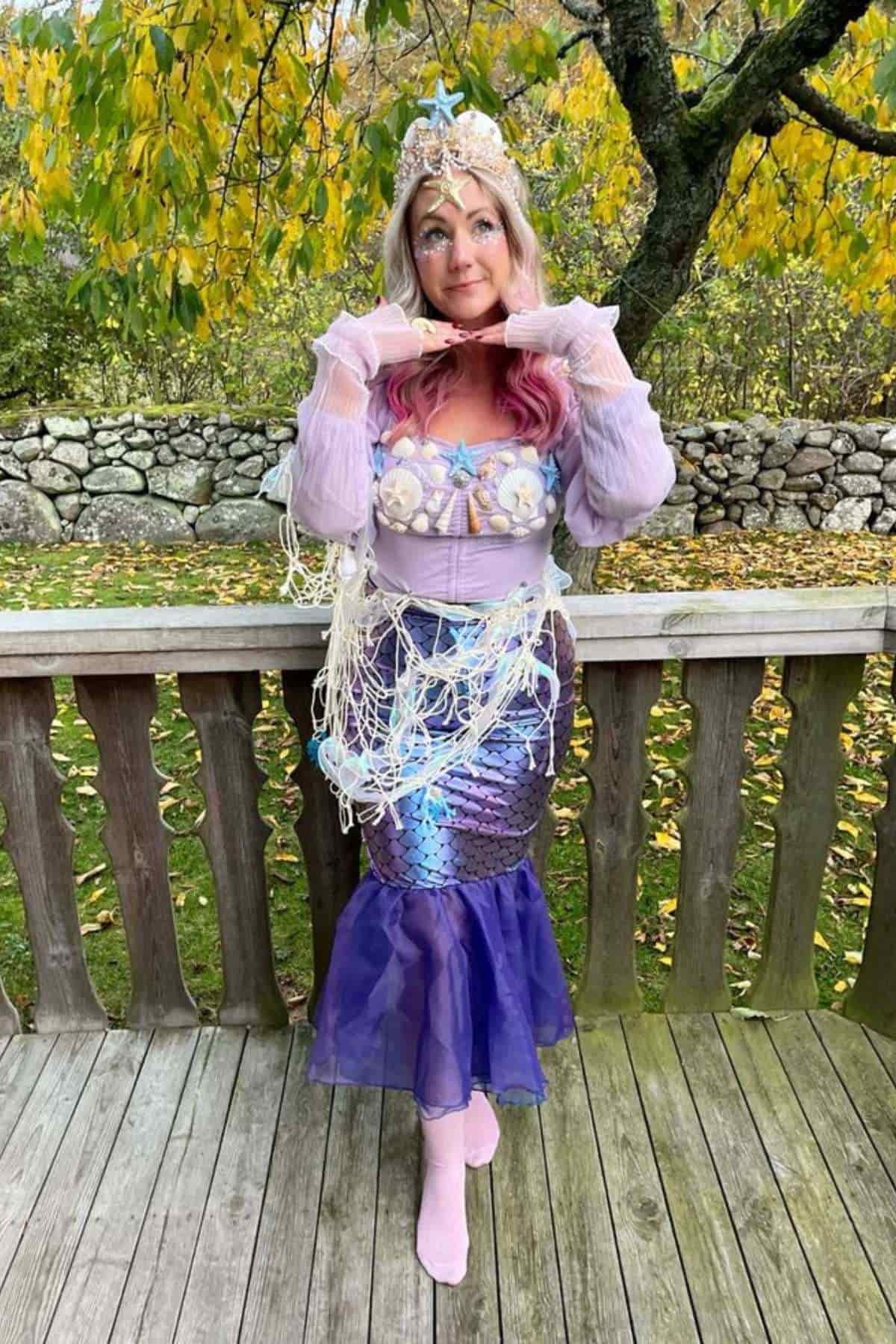 A person in a mermaid costume with a seashell top, shimmery tail, and crown poses outside on a wooden deck, surrounded by autumn trees and a stone wall.
