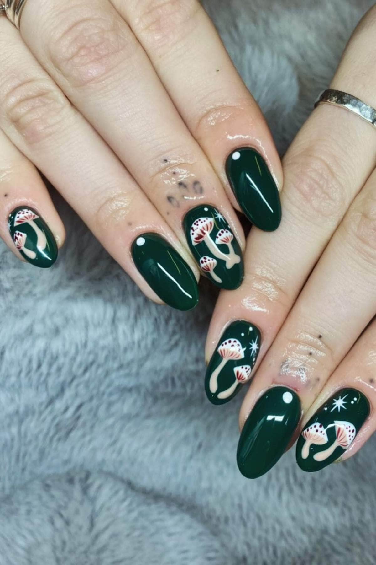 Hands with dark green almond-shaped nails featuring mushroom and star designs on some fingers, placed on a gray textured surface.