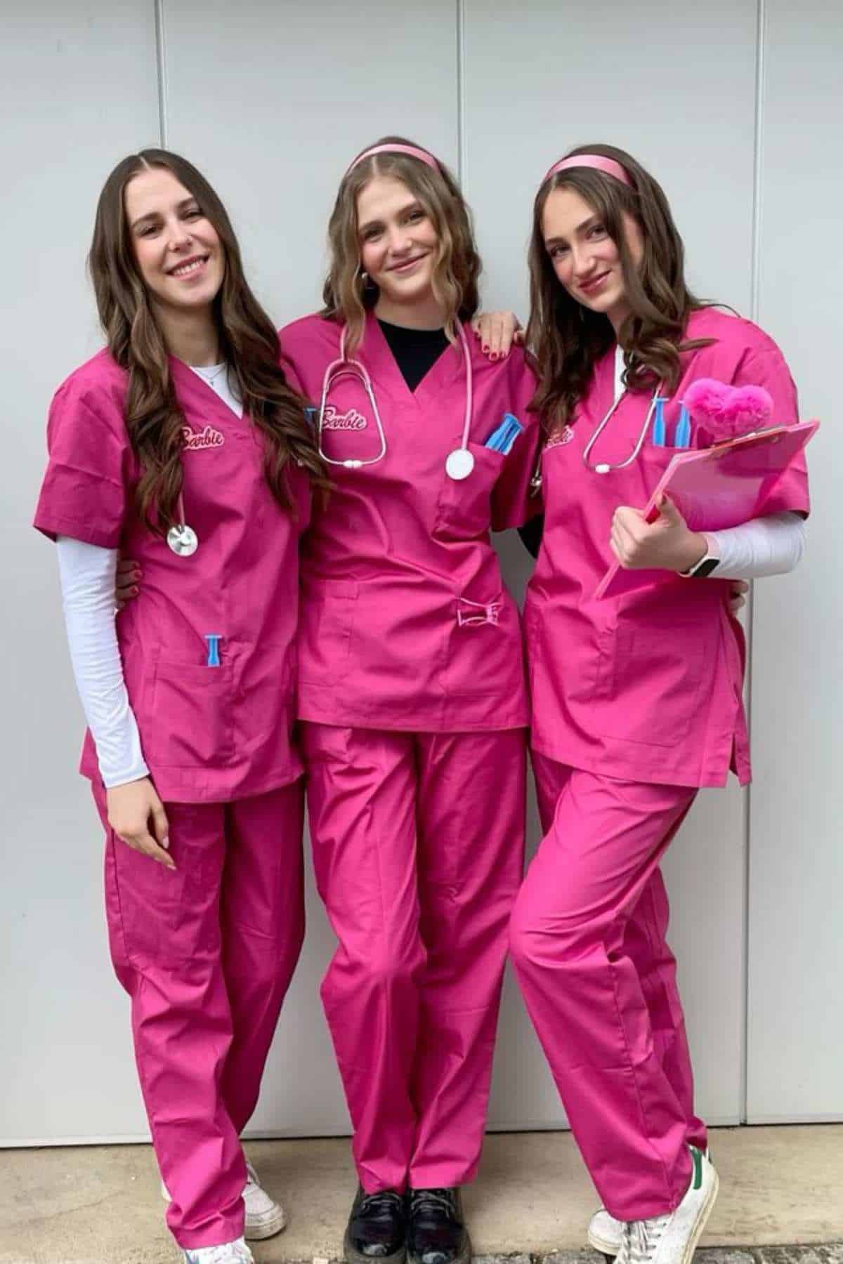 Three people wearing bright pink medical scrubs and headbands stand together, smiling, in front of a plain white wall. One holds a clipboard with pink papers and a pen.