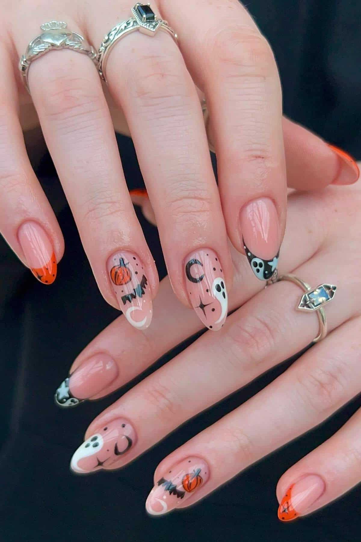 Hands with long almond-shaped nails featuring Halloween-themed nail art, including pumpkins, bats, moons, and skulls on nude and white polish with orange and black details.