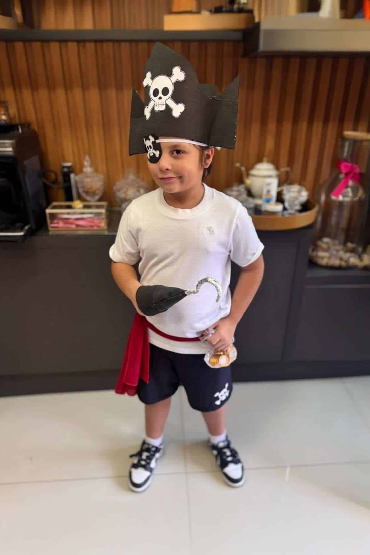 Young boy in a pirate costume, wearing a black hat with a skull and crossbones, holding a hook prop and sword, standing indoors in front of a shelf.