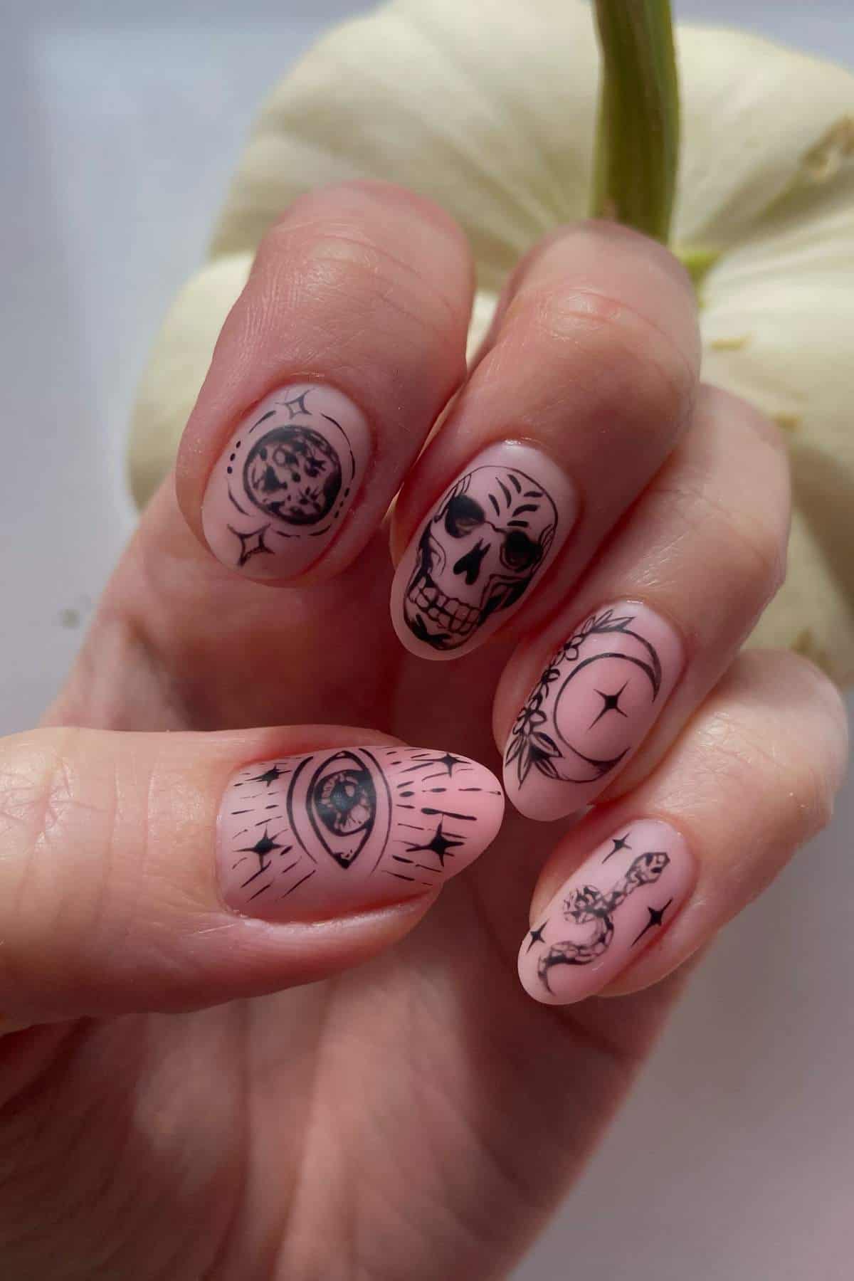 A hand with pale pink nails featuring black tattoo-style art, including a skull, eye, moon, snake, and stars, held in front of a white pumpkin.