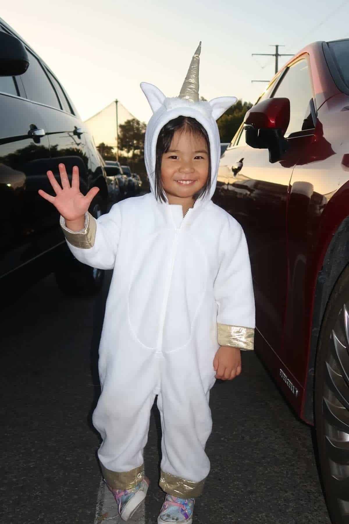 Young child in a white unicorn costume with gold accents stands between parked cars, smiling and waving at the camera.