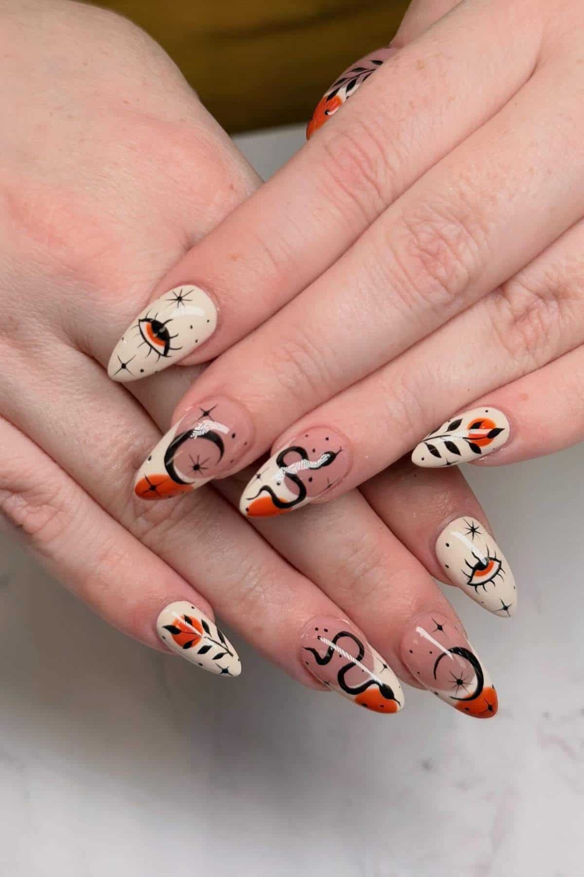 Close-up of hands with almond-shaped nails, featuring abstract black, orange, and white designs with eyes, leaves, and star-like accents on a light and nude base.