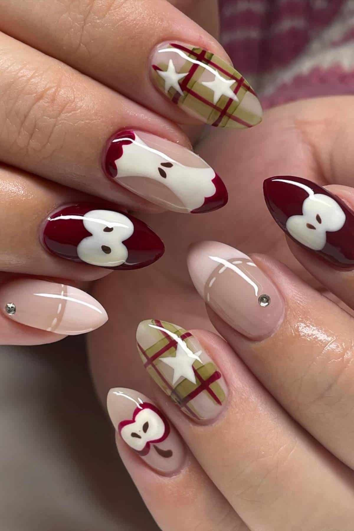 Close-up of hands with almond-shaped nails featuring burgundy, plaid, apple, and abstract designs, along with small rhinestone accents on some nails.