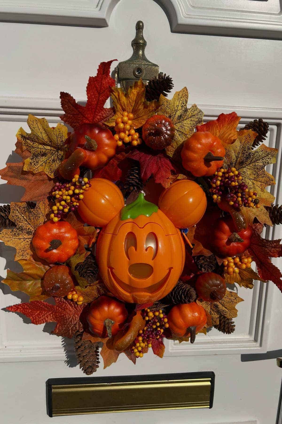 A fall wreath with orange leaves, pumpkins, pinecones, berries, and a plastic Mickey Mouse jack-o'-lantern is hanging on a white door.