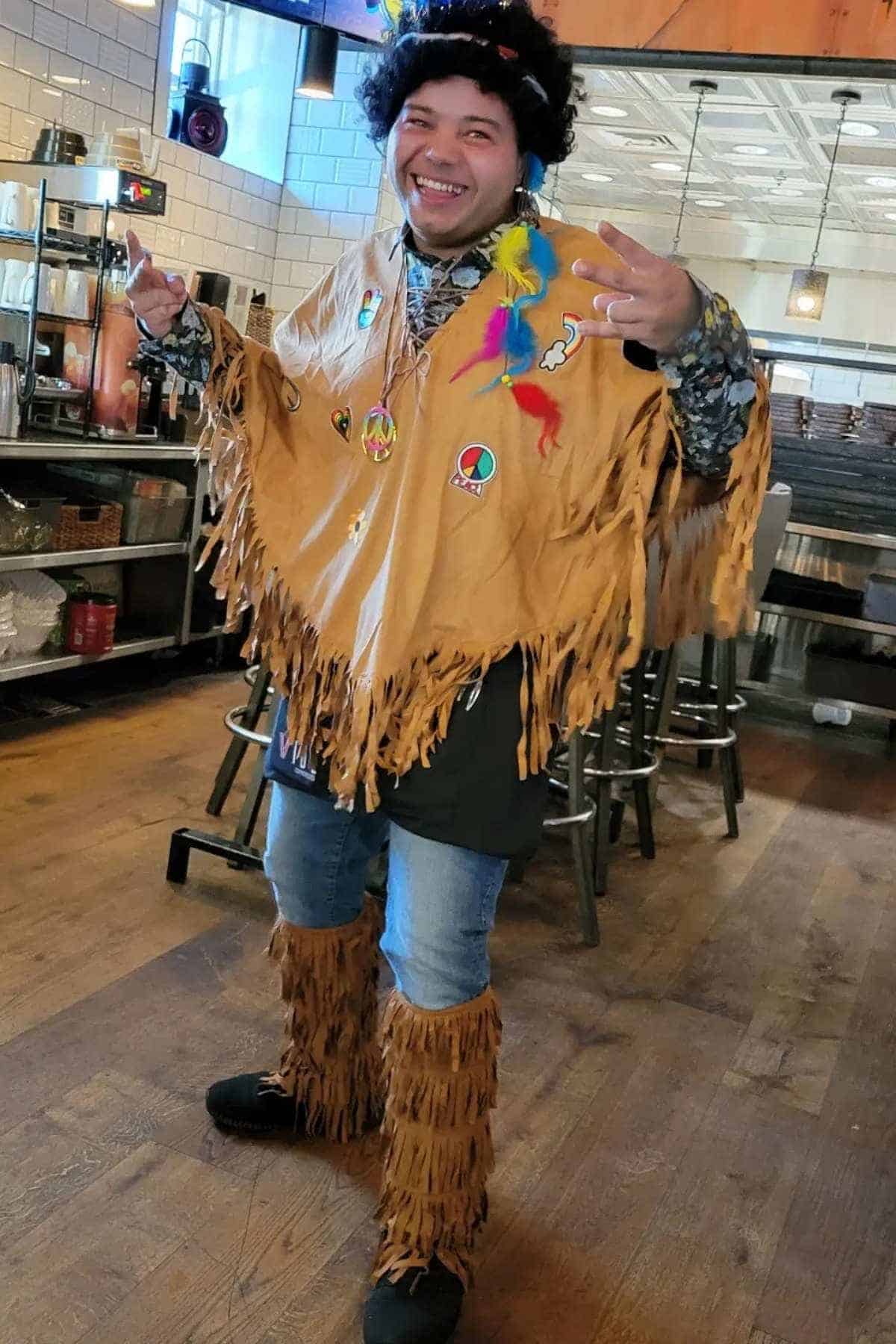 A person indoors wears a fringed tan poncho, matching leg coverings, a black wig, and colorful feather accessories while smiling and making a peace sign gesture.