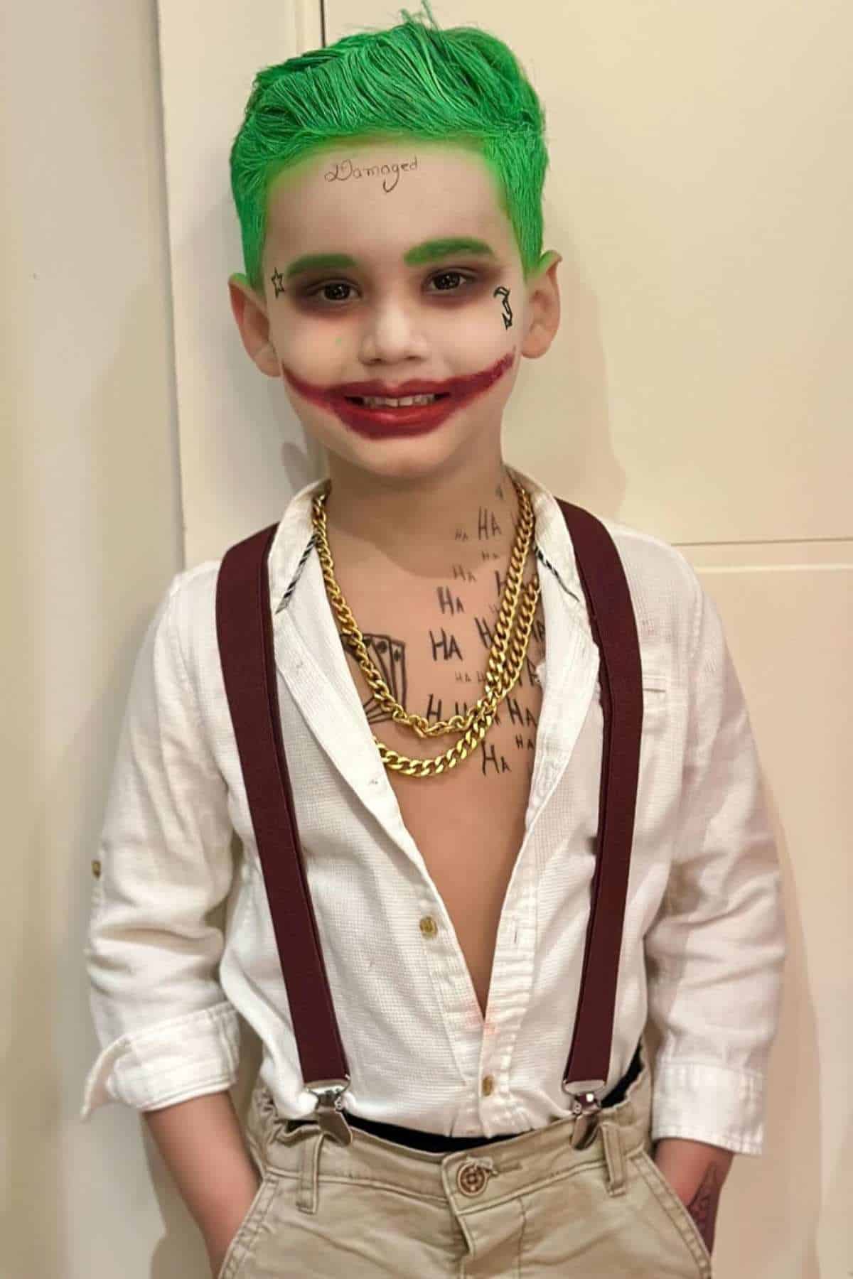 Young child dressed as the Joker with green hair, white shirt, suspenders, gold chains, face paint, and fake tattoos.