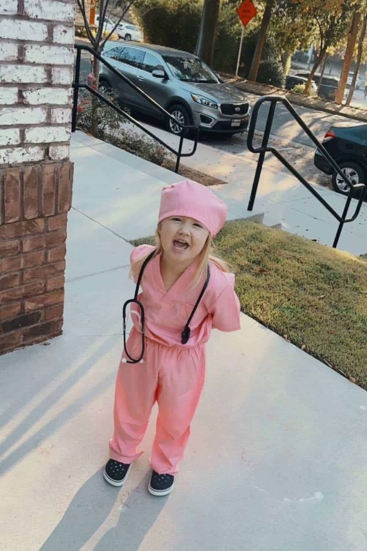 Young child in pink scrubs and a stethoscope stands on a sidewalk outdoors, looking up with mouth open.