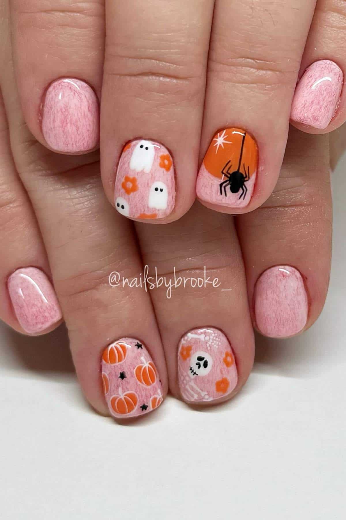 Short nails with pink polish, featuring Halloween-themed designs: ghosts, pumpkins, a black spider, a skull, and white spider webs.