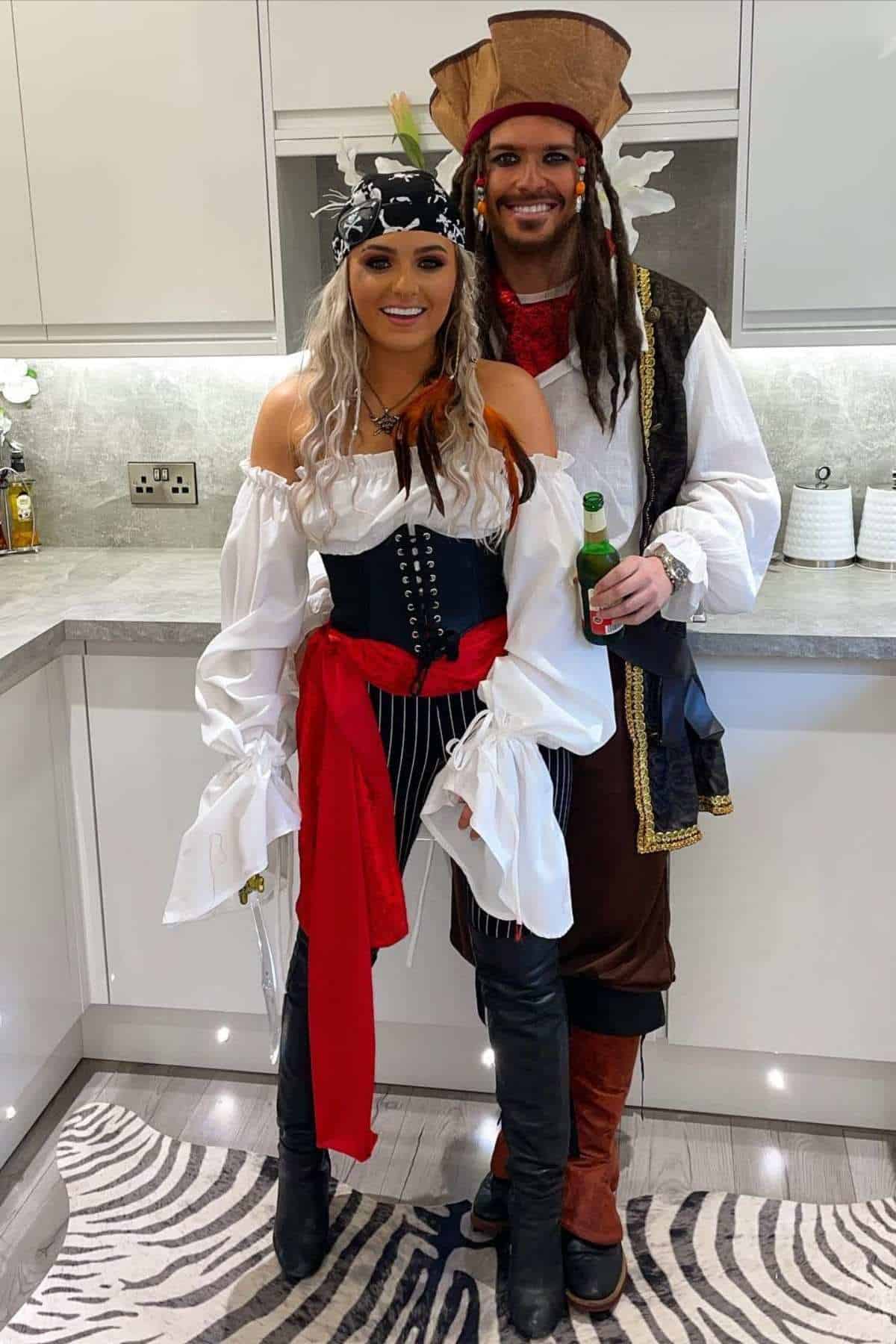 Two adults in pirate costumes stand together in a modern kitchen. The woman wears a corset and striped pants; the man wears a tricorn hat and holds a bottle.
