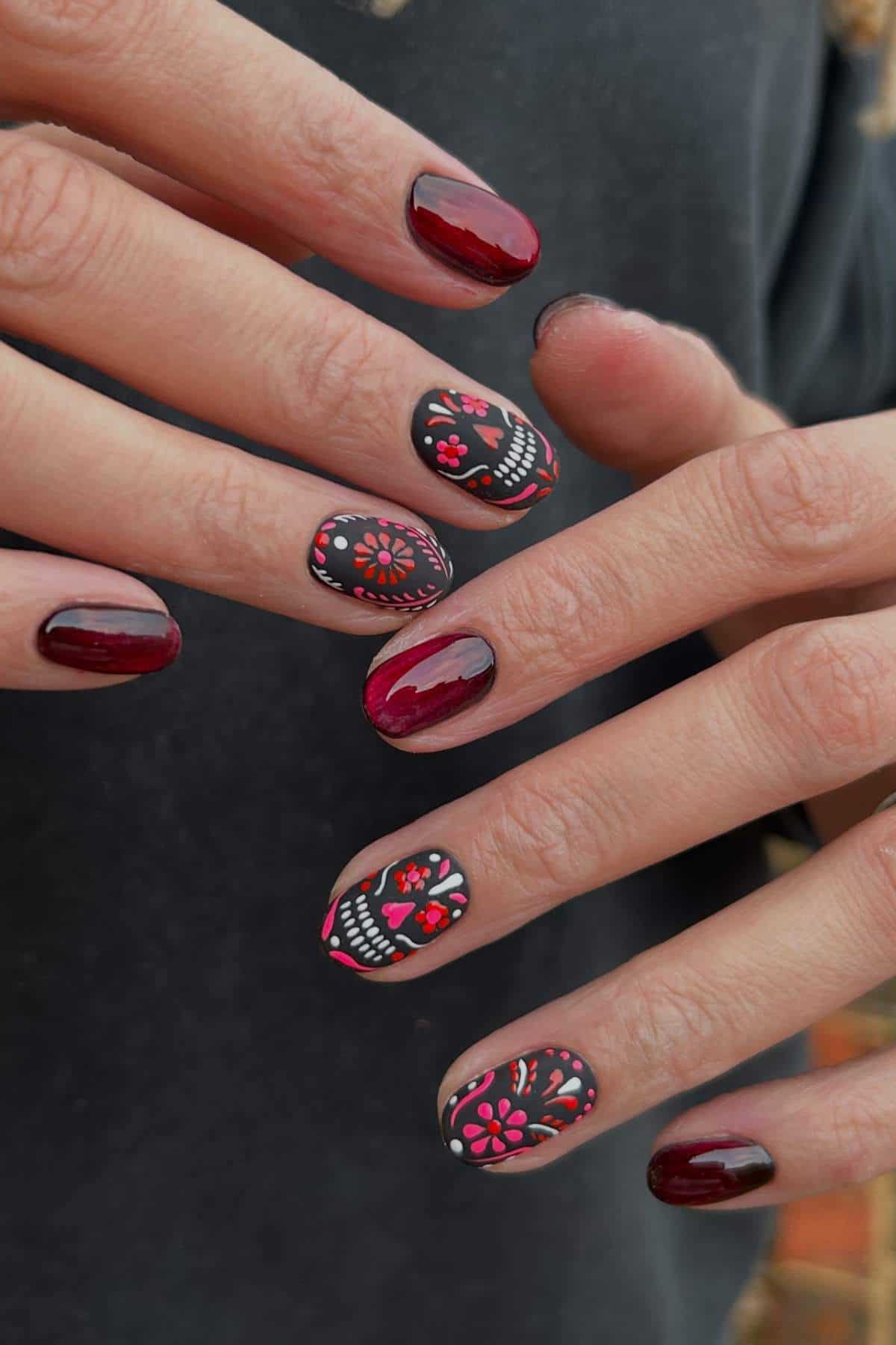 Hands with dark red and black nail polish; some nails feature detailed sugar skull and floral designs in pink and red.