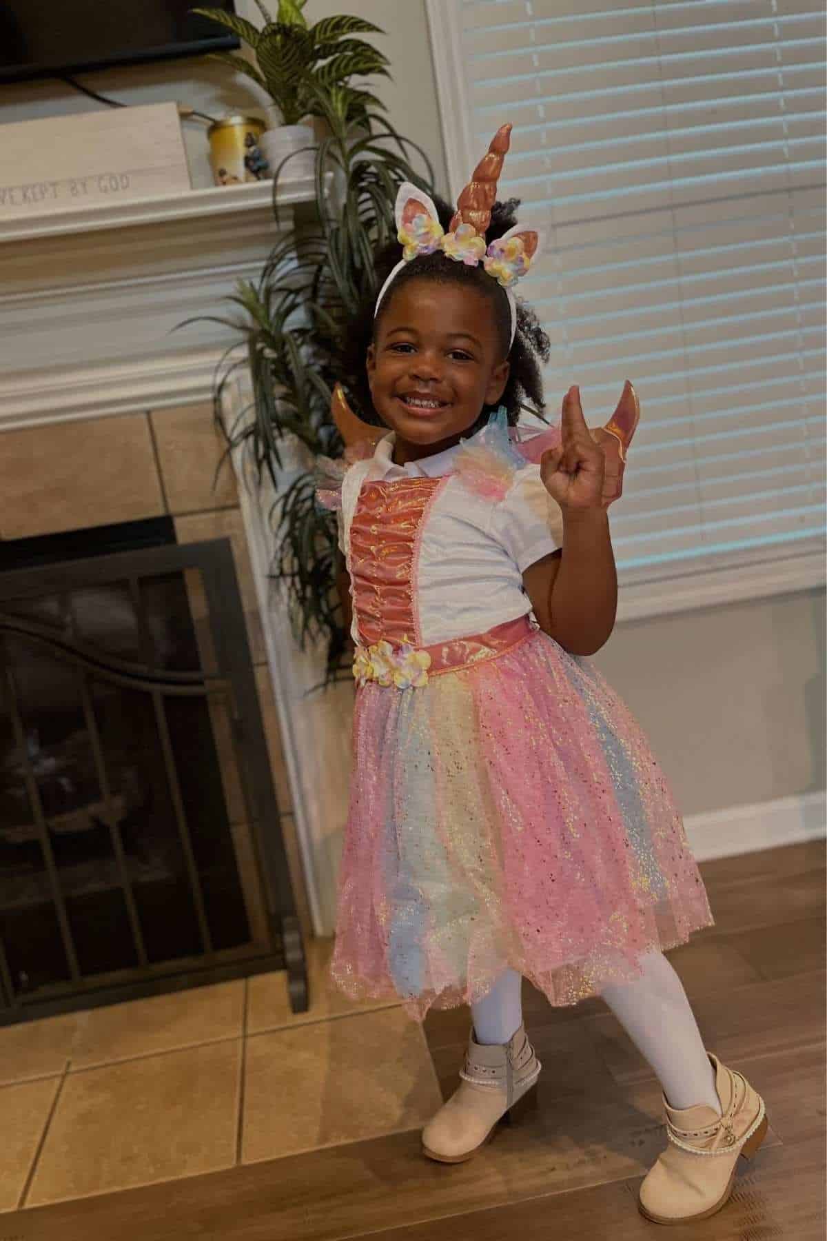 A young child smiles and poses indoors wearing a pastel unicorn costume with a horn headband, wings, and a colorful dress.