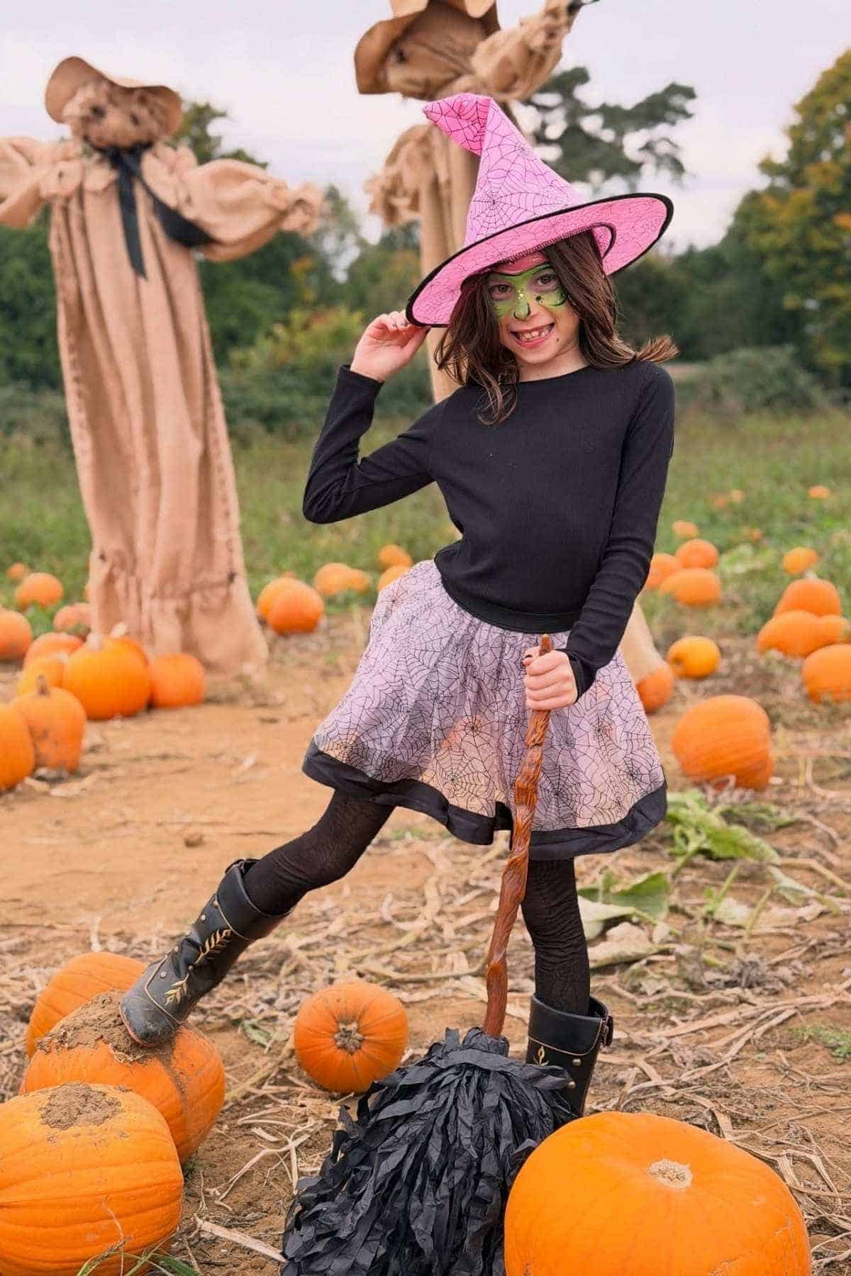 A child in a witch costume stands on a pumpkin in a pumpkin patch, holding a broomstick, with scarecrows and scattered pumpkins in the background.