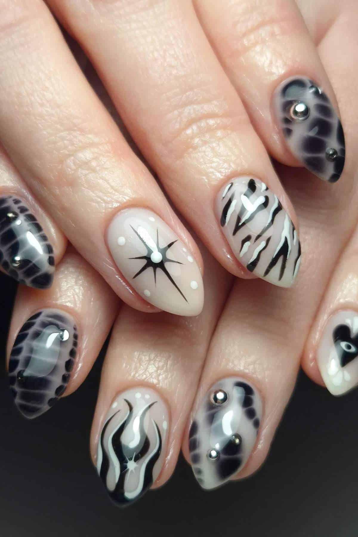 Hands with almond-shaped nails featuring black and white abstract designs, dots, and small gem accents on a neutral background.