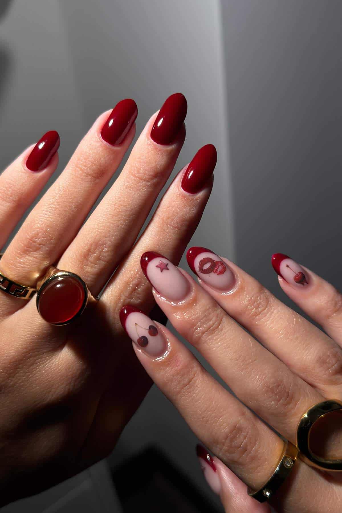 A person displays manicured hands with deep red nails; some nails feature clear polish with red abstract designs and symbols. The person wears gold rings on their fingers.