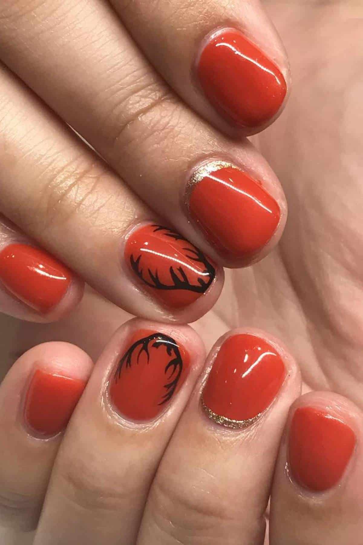 Close-up of hands with short, glossy, orange-red nails; one nail features an antler design, and one has gold detailing near the cuticle.