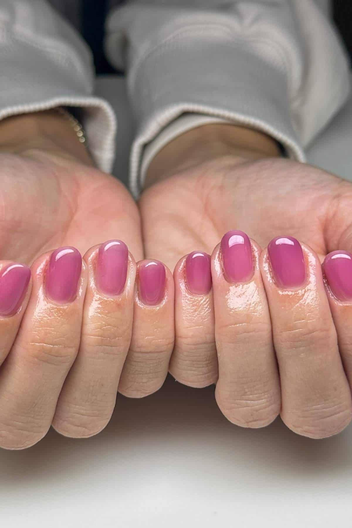 Close-up of two hands showing short, well-manicured nails painted with glossy pink nail polish against a light background.