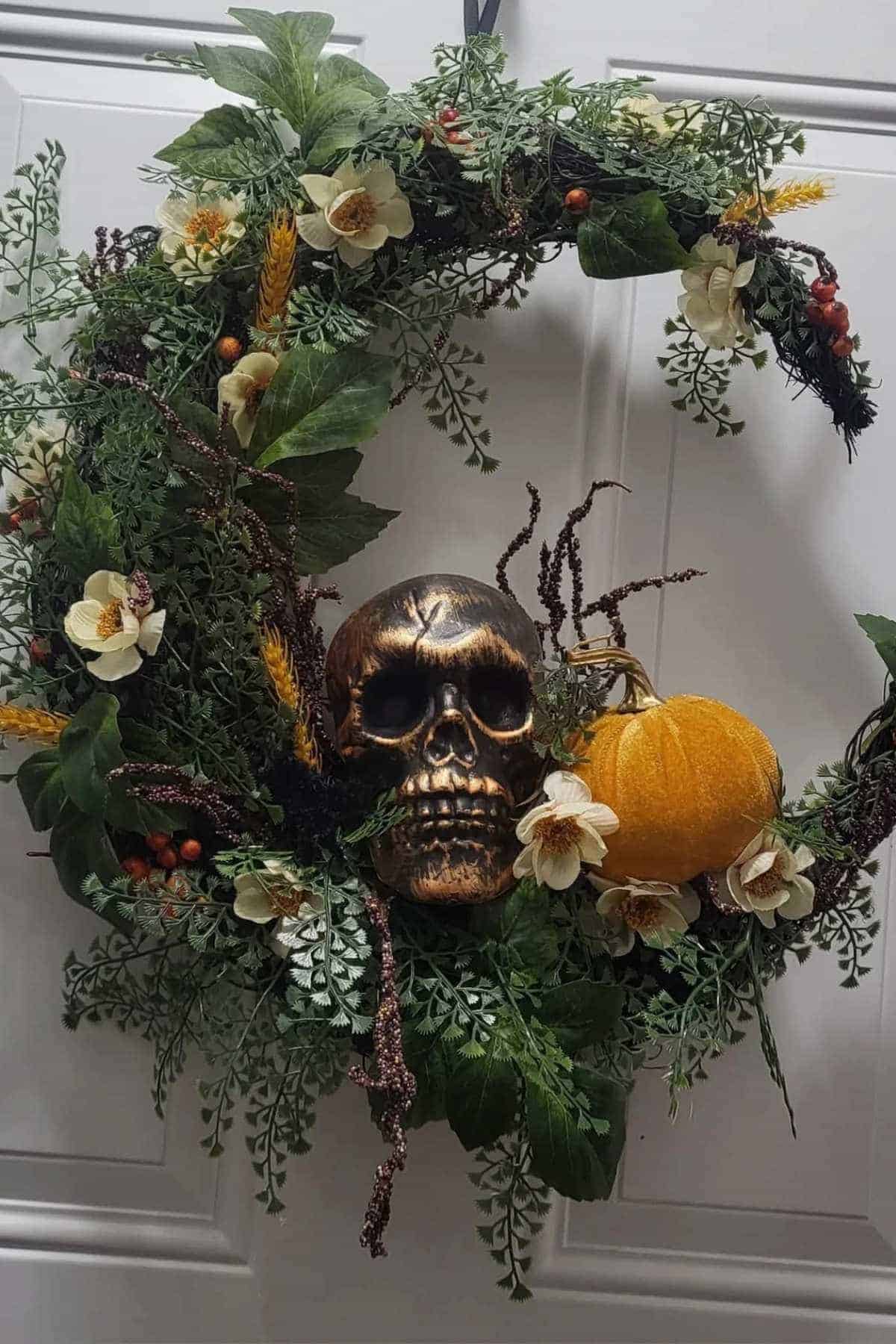 A decorative wreath with green leaves, white flowers, a gold skull, orange pumpkin, and red berries, hanging on a white door.