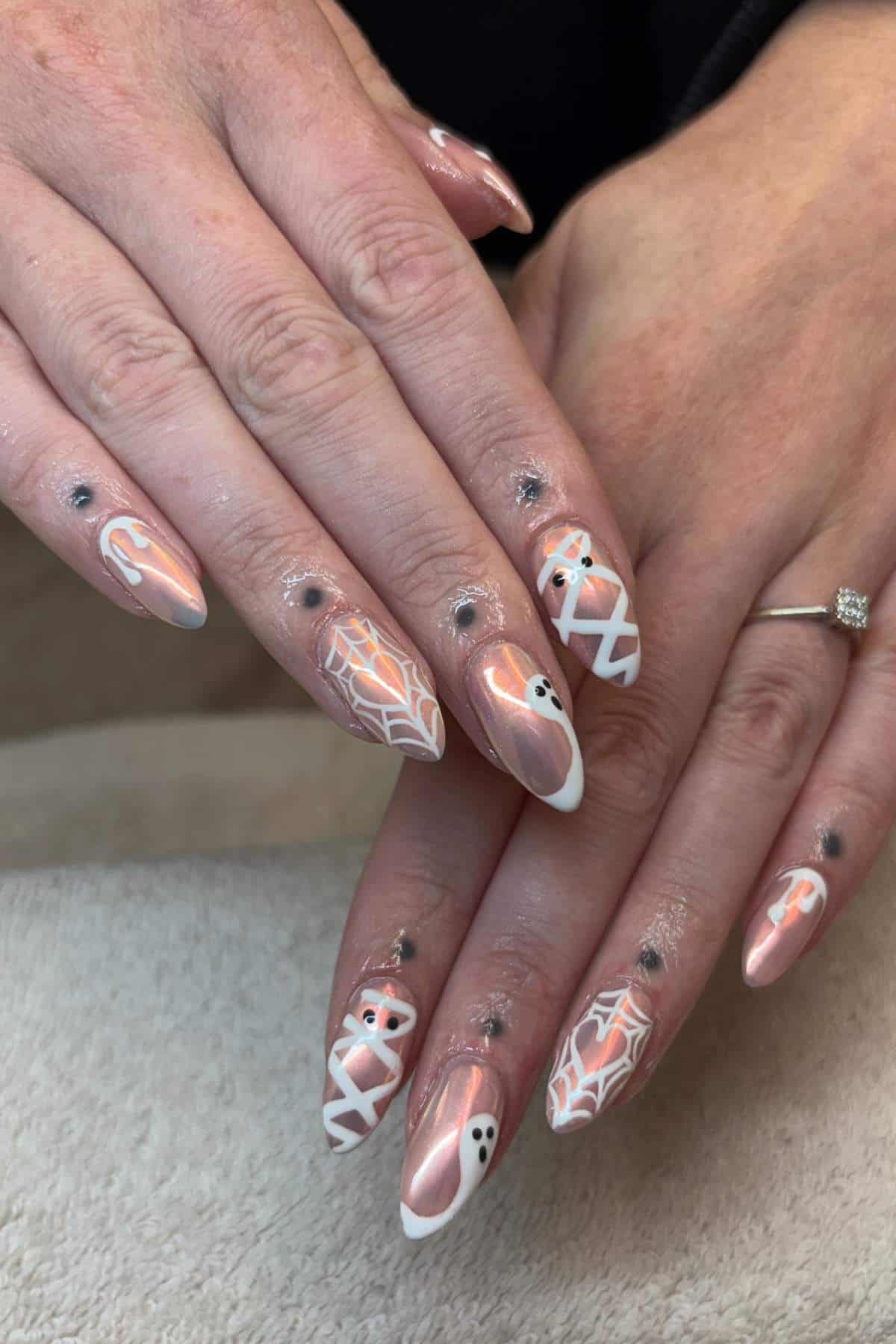 Hands with long, almond-shaped nails featuring Halloween-themed nail art such as webs, ghosts, and eyes, along with small black gem accents on fingers.