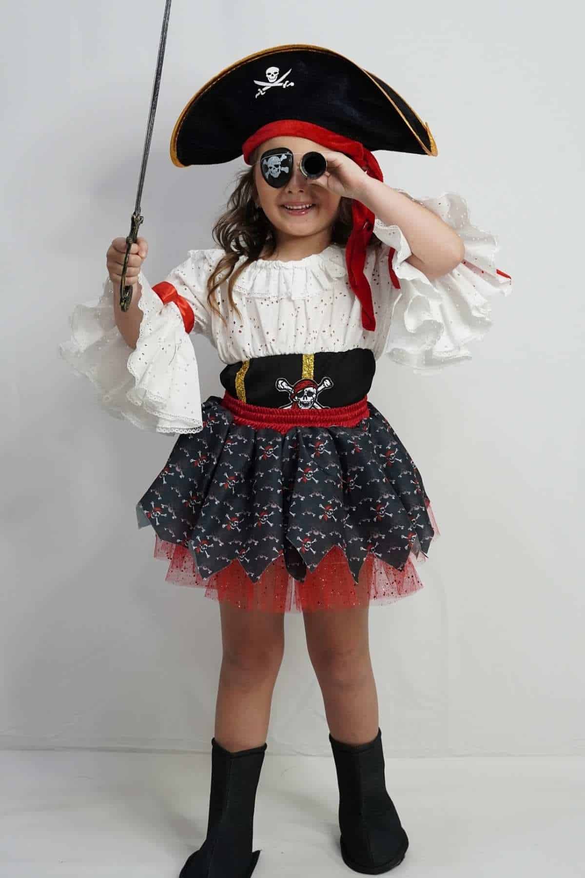 Child dressed as a pirate holds a sword and salutes with one hand. She wears a pirate hat, eye patch, white shirt, black skirt, red sash, and black boots against a plain background.