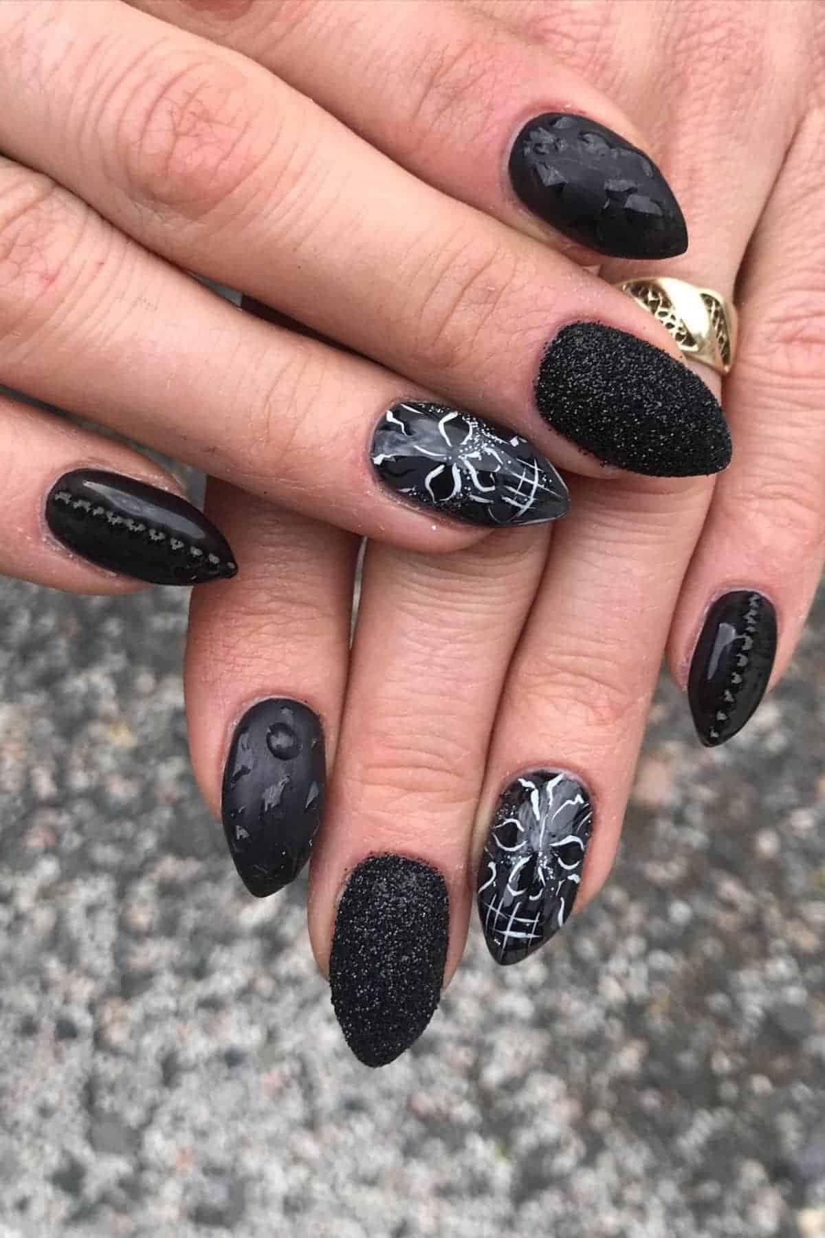 Hands with black, pointed nails featuring various textured designs, including matte, glossy, beaded, and intricate white line art on two nails, shown against a speckled background.