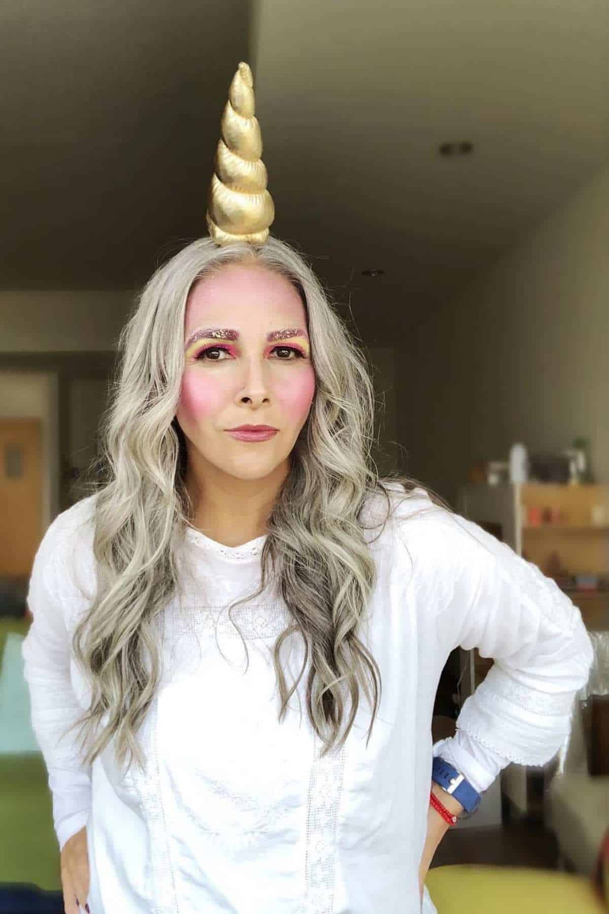 Person with long gray hair, wearing a white top, gold unicorn horn headband, and colorful makeup, standing indoors with shelves and various items in the background.