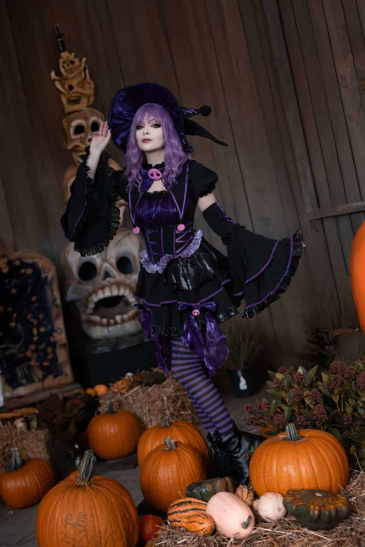 Person in a purple witch costume stands among pumpkins and gourds, with Halloween-themed decorations and wooden planks in the background.