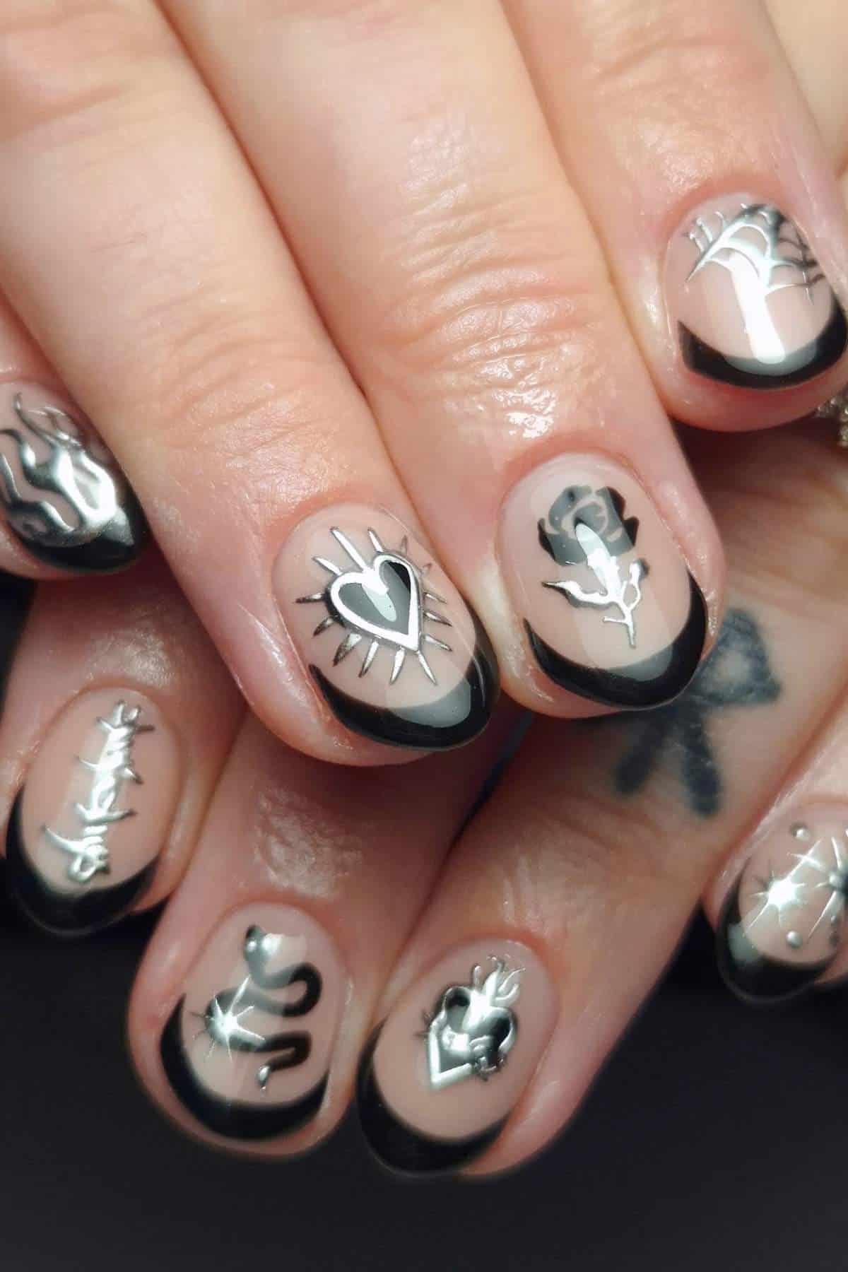 Closeup of hands with nude nails featuring black French tips and various black and white tattoo-inspired designs, including hearts, flames, webs, and snakes.