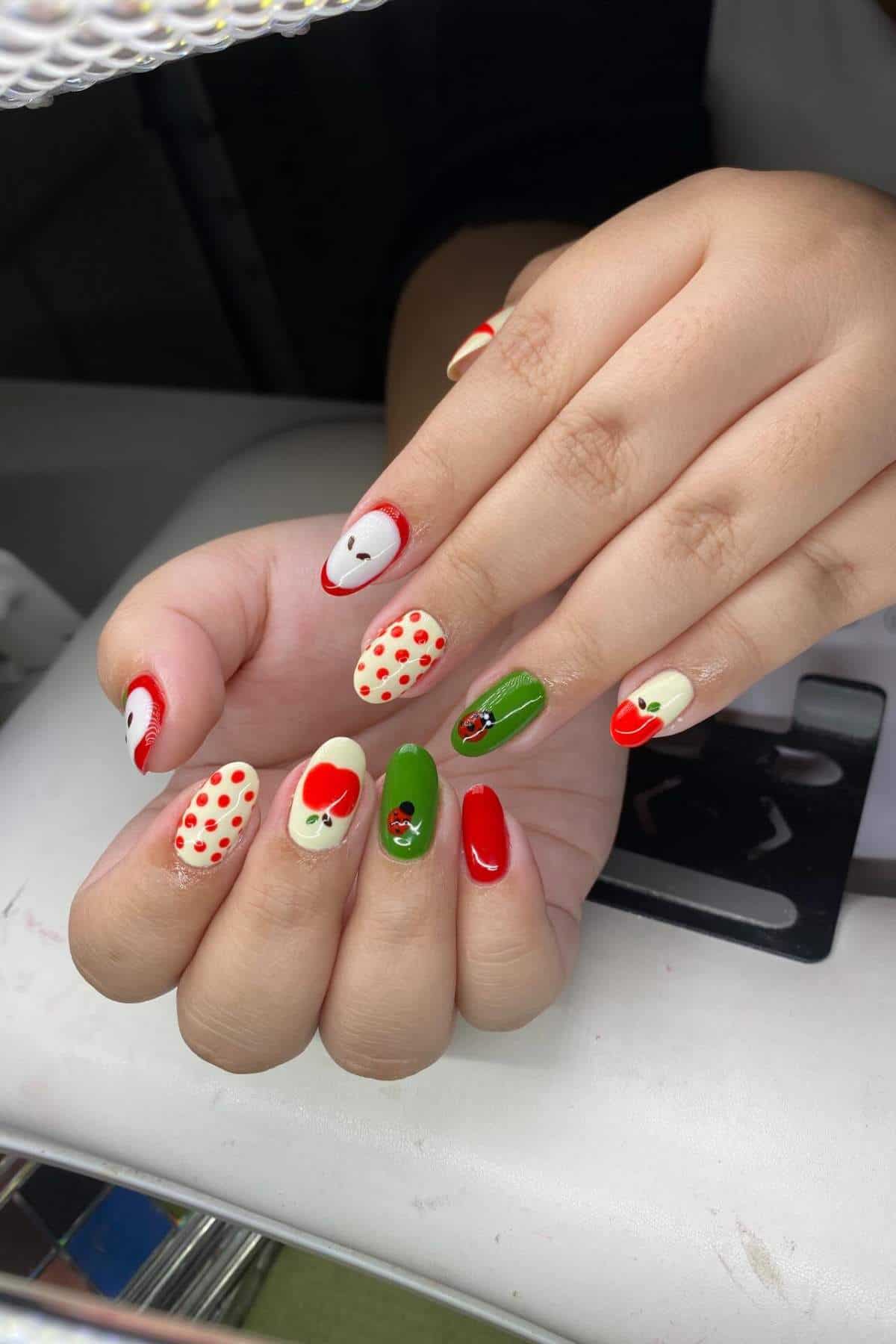 A hand with long, almond-shaped nails painted in red, green, and cream, featuring polka dots, apple designs, and small decorative embellishments.