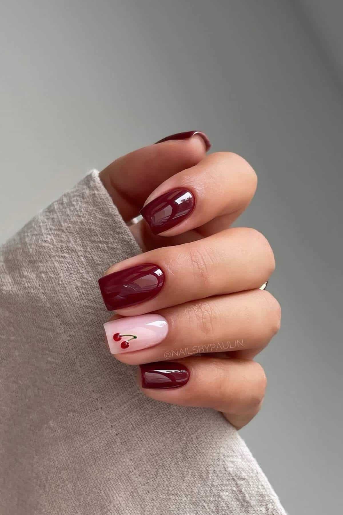 A hand with short, square-shaped nails painted dark red, except the ring finger is pale pink with a small red cherry design.