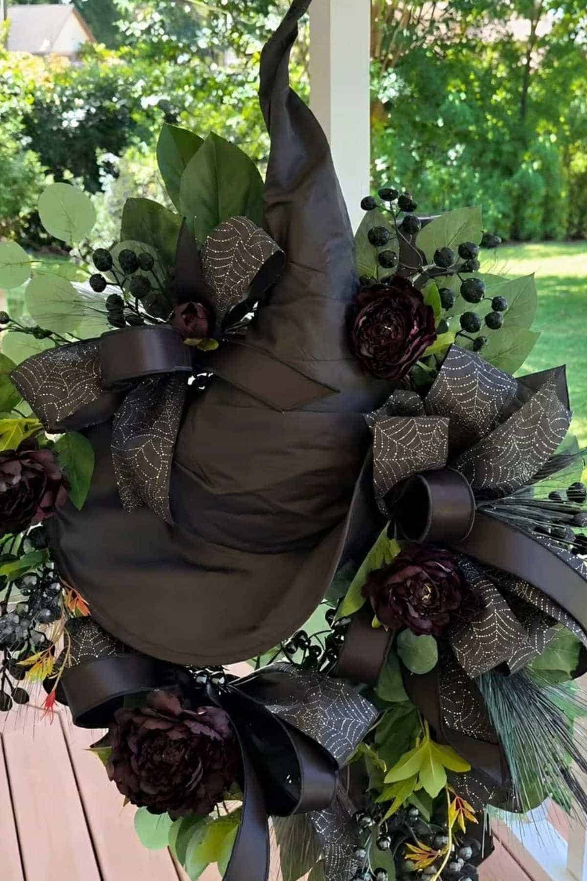 A wreath featuring a black witch hat, dark flowers, black ribbons with spiderweb designs, and green foliage, hanging outside on a porch.