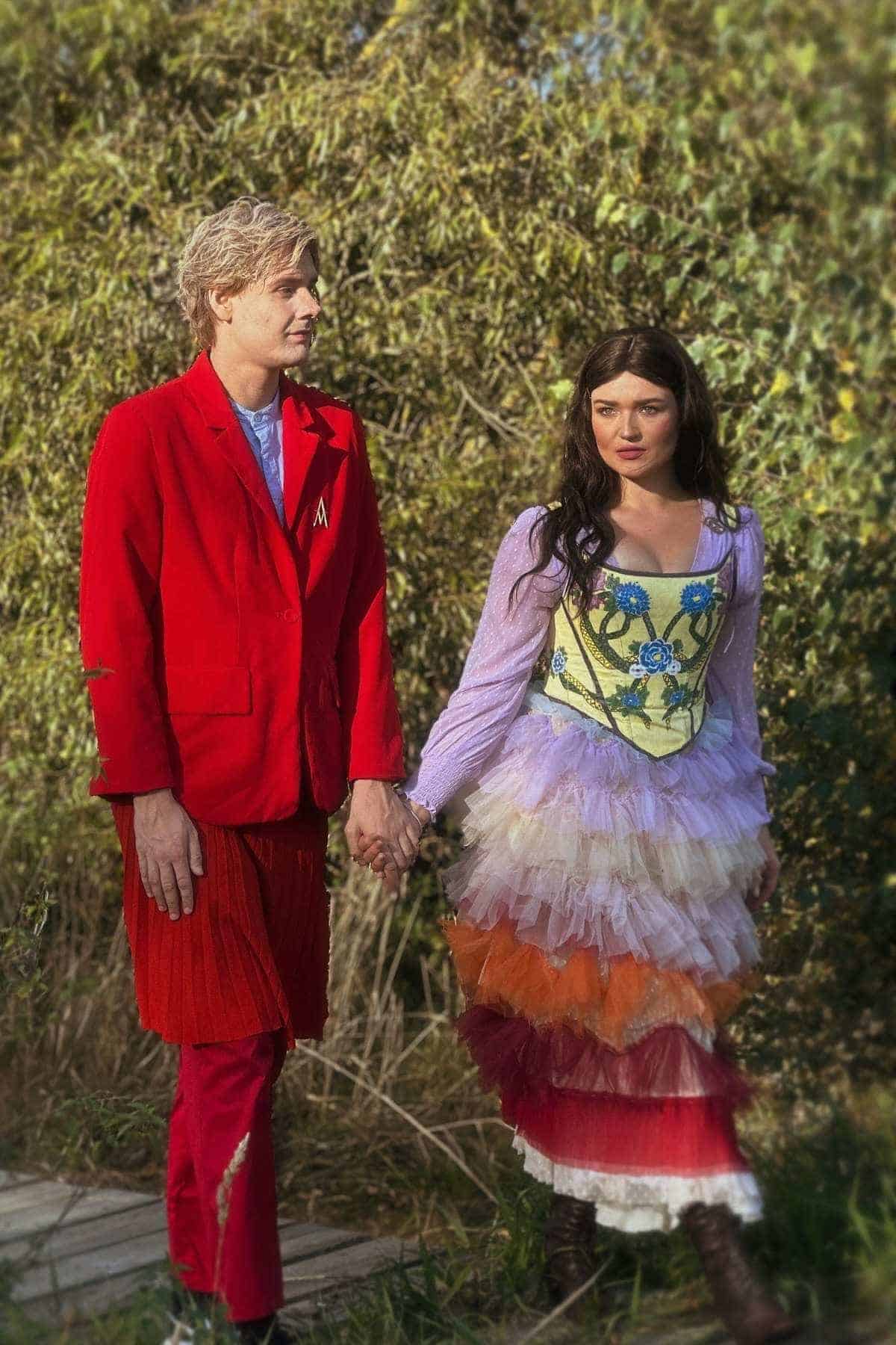 Two people hold hands outdoors; one wears a bright red suit, the other wears a layered dress with floral and colorful ruffle details. Trees and greenery are in the background.