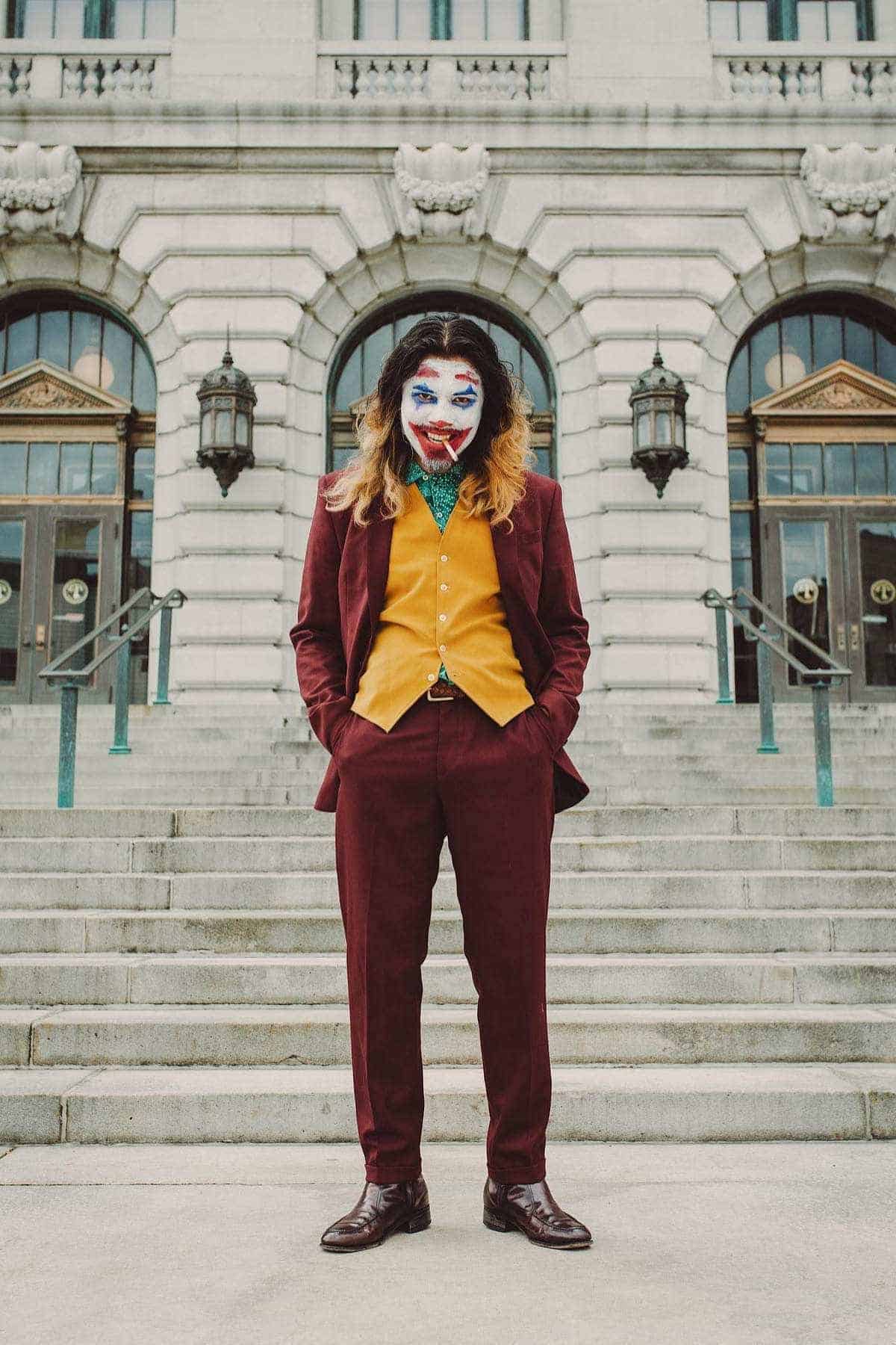 Person in a maroon suit and gold vest stands on stone steps outside a building, wearing clown makeup and a green shirt, facing forward with hands in pockets.