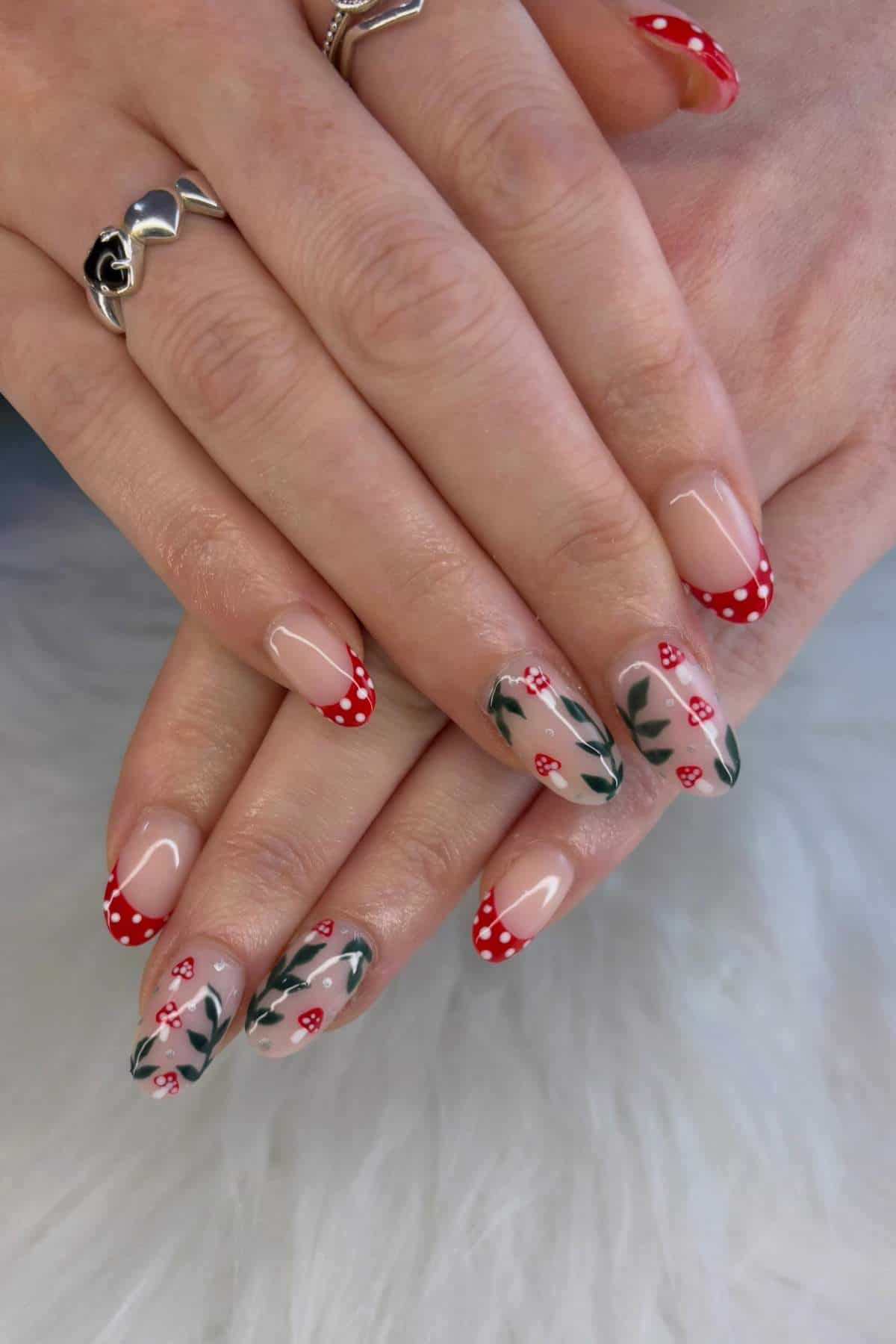 Hands with manicured nails featuring red and white mushroom designs, green leafy accents, and a nude base. Rings are worn on two fingers.