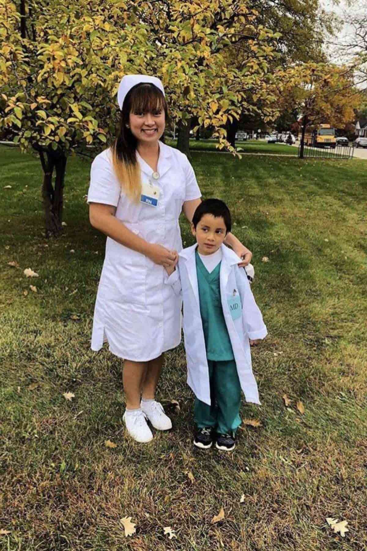A woman in a nurse uniform stands on grass next to a young boy dressed in a white coat and scrubs. Trees with autumn leaves appear in the background.