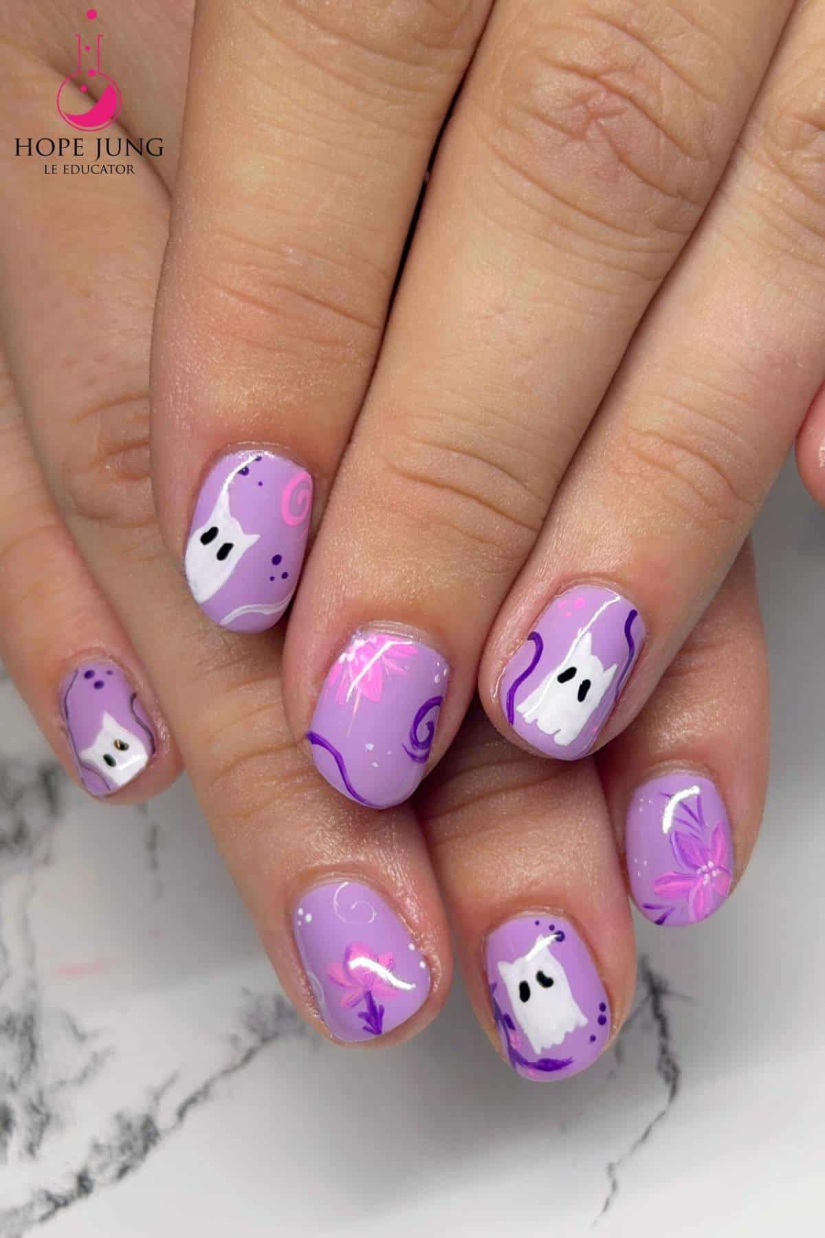 Close-up of hands with short nails painted purple, featuring white ghost designs, black dots, and pink and purple swirls.