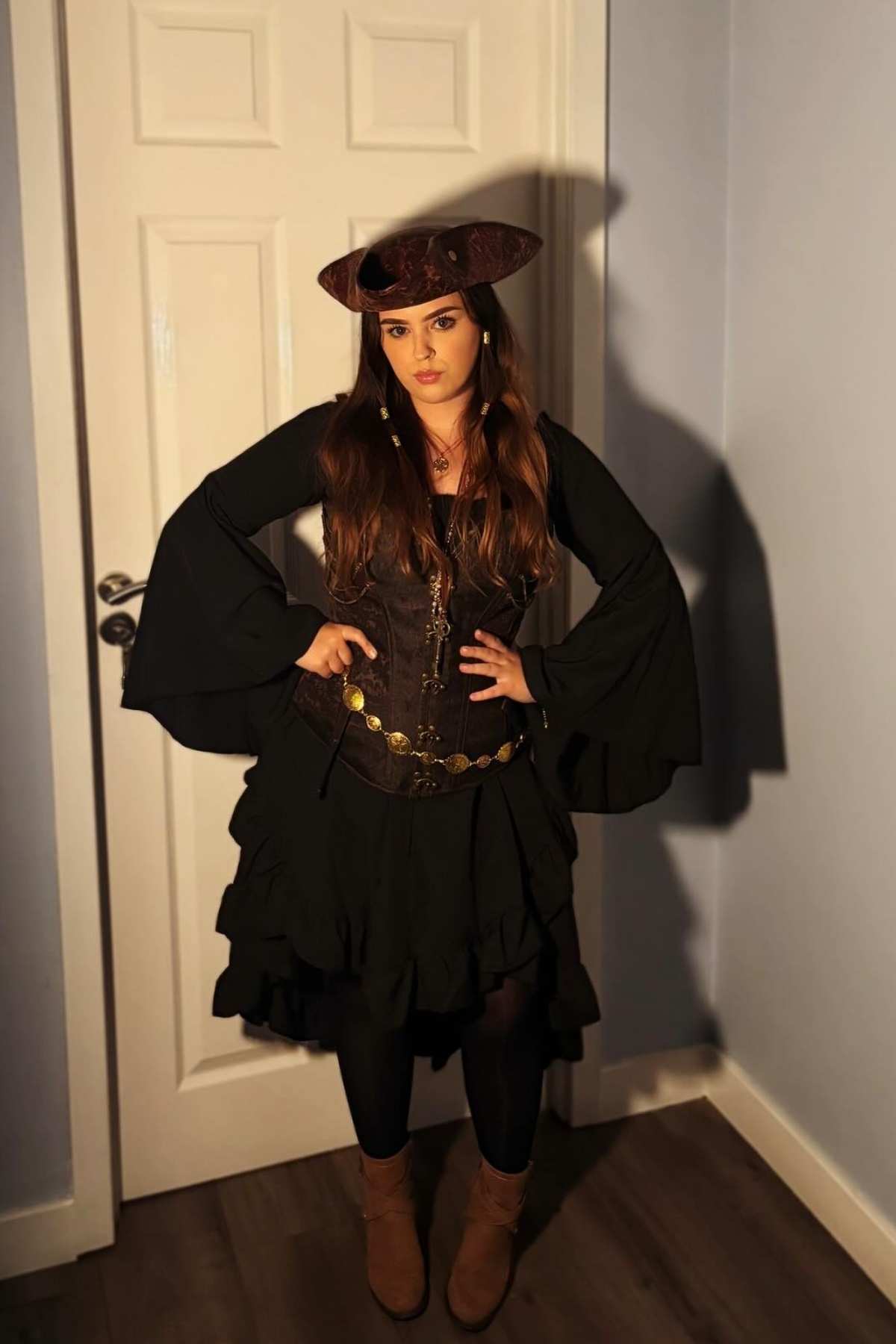 Woman stands indoors wearing a dark pirate costume with flared sleeves, corset, ruffled skirt, brown boots, and a pirate hat, posing with hands on hips against a white door.