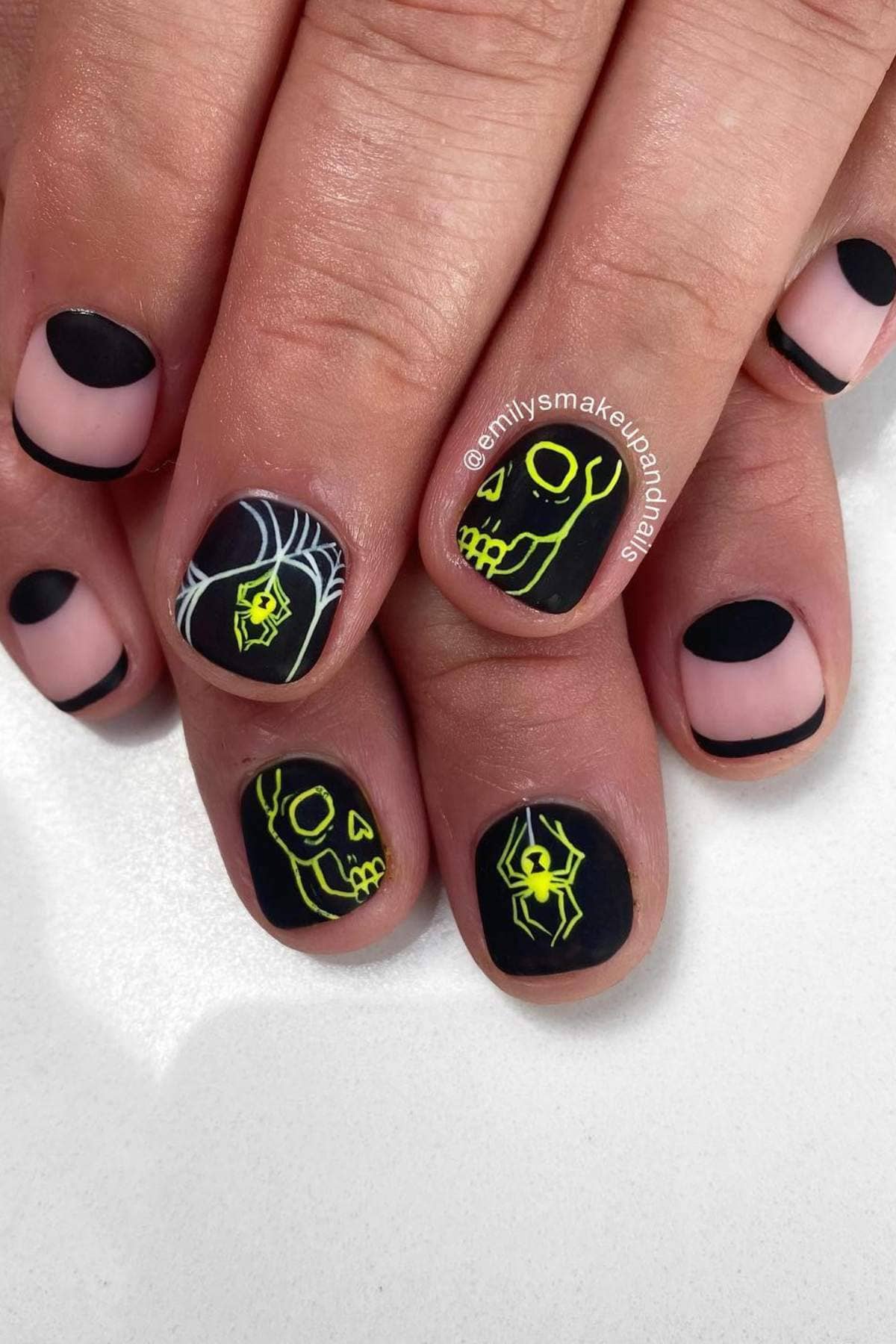 Fingernails with black tips and Halloween designs, including neon green spiders, spider webs, and skulls on a matte black background.