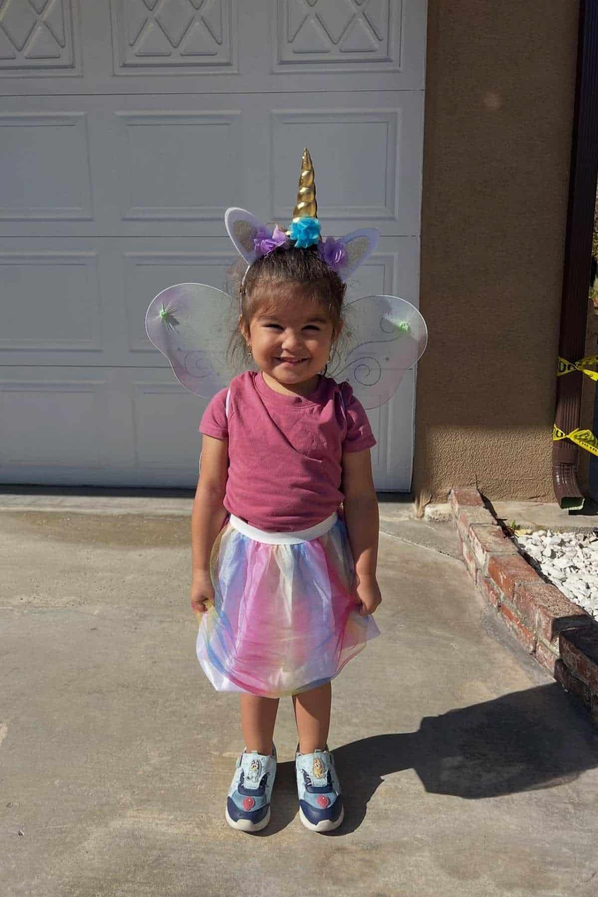 Young child stands outside in front of a garage, wearing a unicorn headband, wings, a pink shirt, multicolored tutu, and sneakers, smiling at the camera on a sunny day.