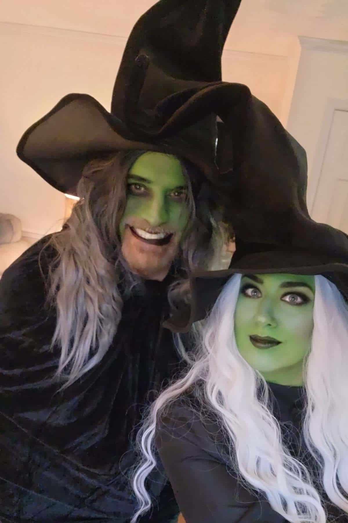 Two people wearing black witch hats, black capes, and long white wigs with green face paint, posing indoors.