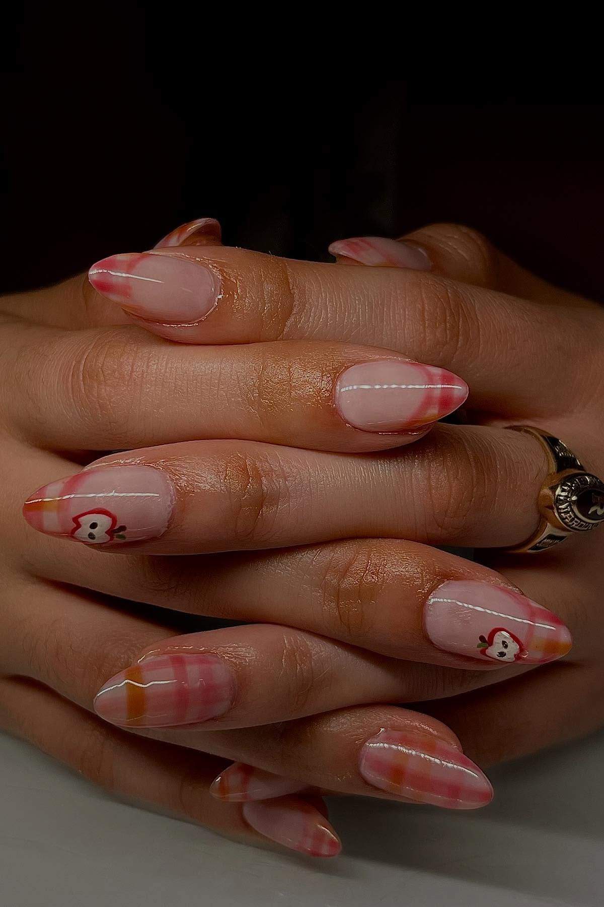 Hands with long, almond-shaped nails featuring a pink and orange plaid design and small heart and bear illustrations on two accent nails.