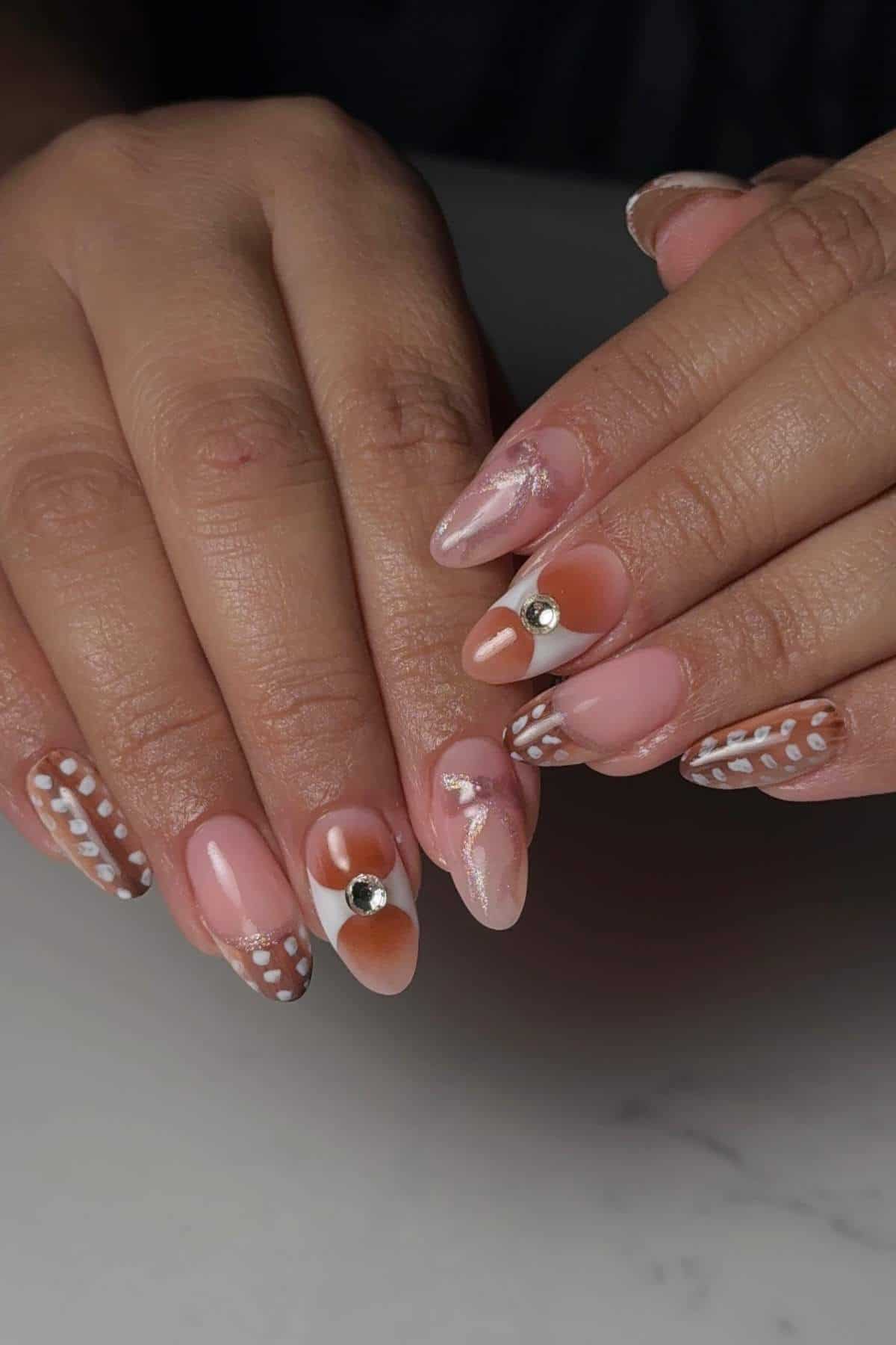 Hands with almond-shaped nails featuring a mix of pink, white, and brown designs, including polka dots, abstract art, and metallic studs.